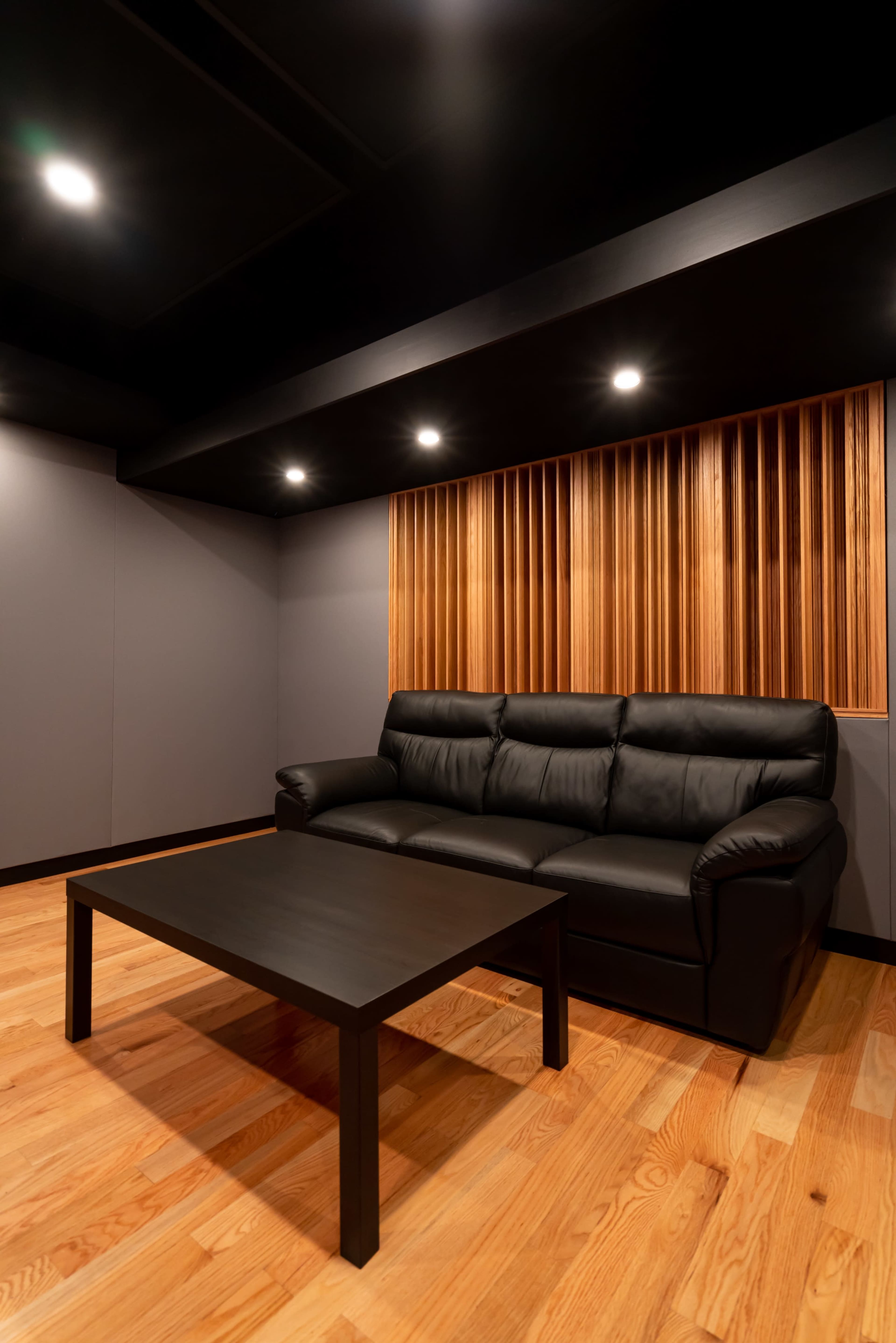 A modern lounge area with a black leather sofa, a black coffee table, and wooden wall accents under bright ceiling lights.