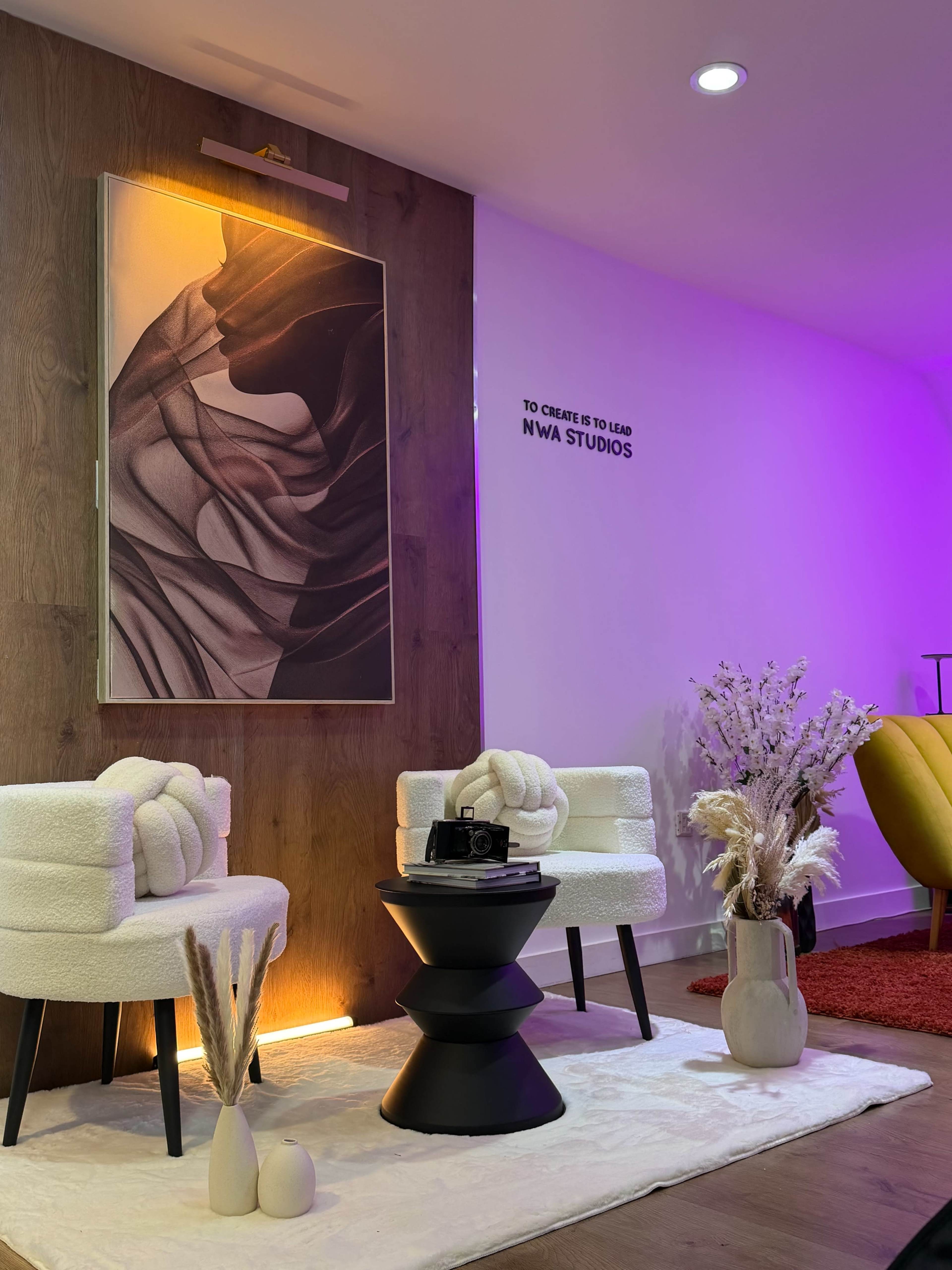 The image shows a modern interior space featuring two white armchairs, a small black table, decorative plants, and a large artwork on the wall, all illuminated with ambient lighting.