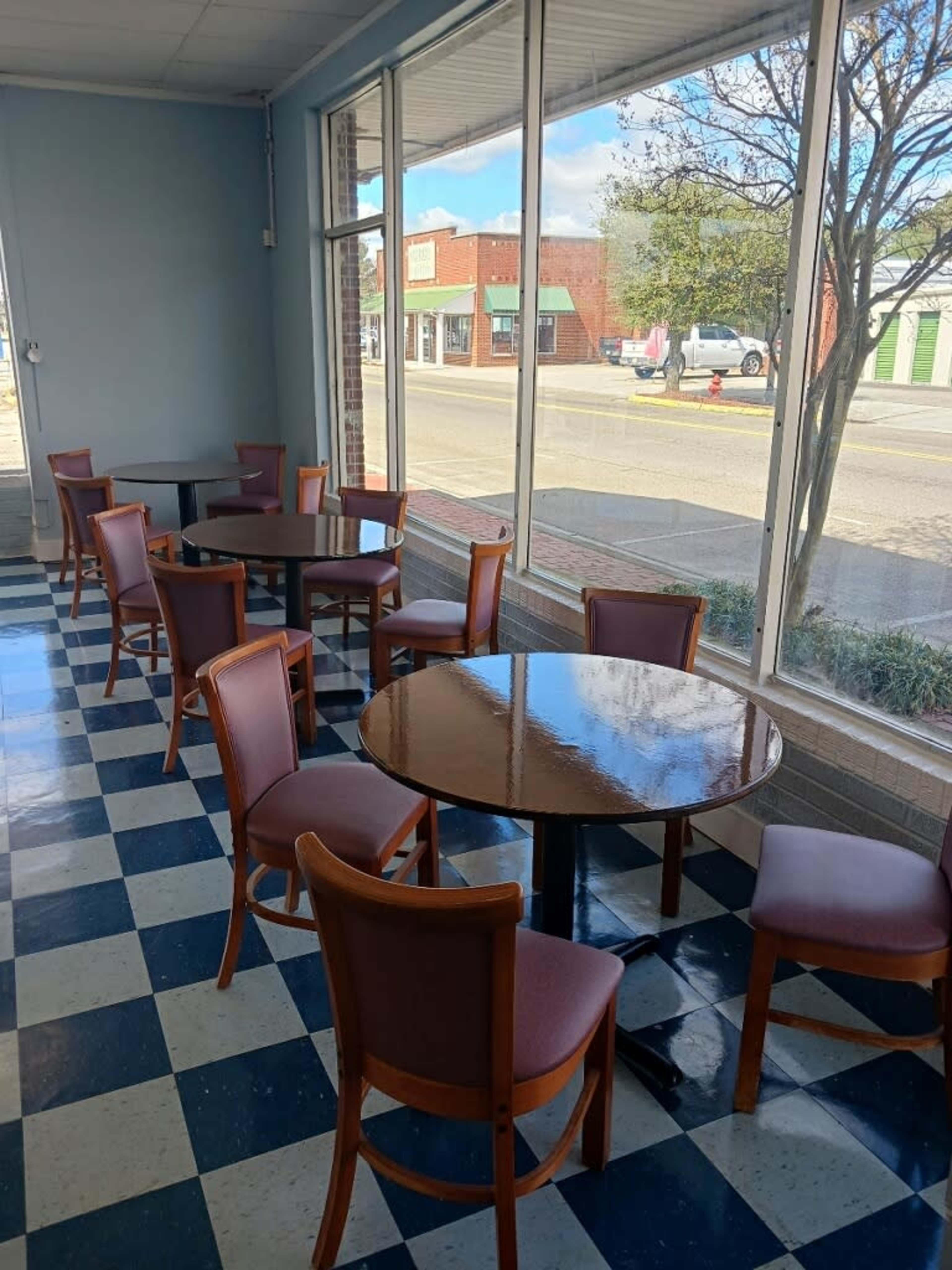 The image shows a small cafe area with several wooden tables and chairs arranged near large windows, overlooking a street.
