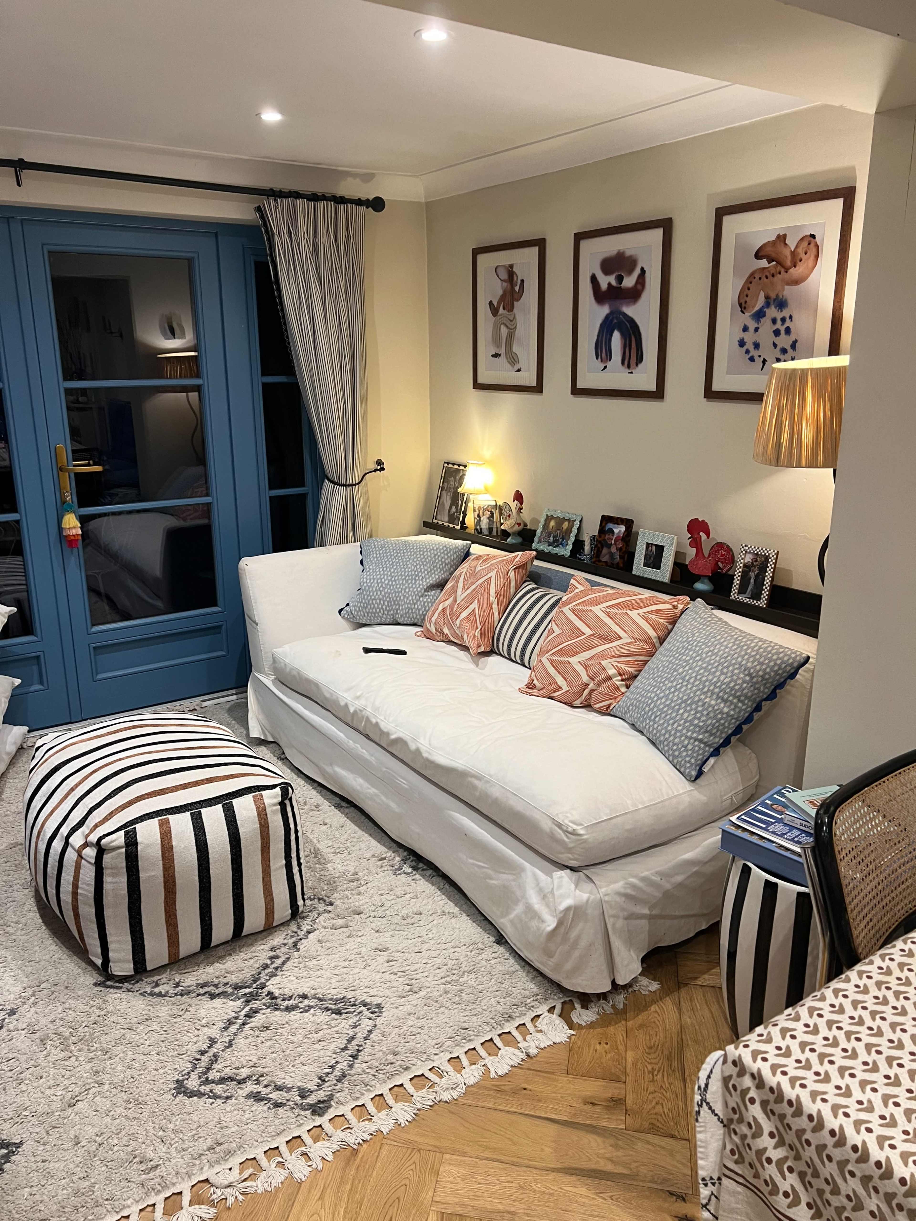 The living room features a white sofa with patterned cushions, a pouf, framed art on the walls, and a lamp next to a blue door leading to another room.
