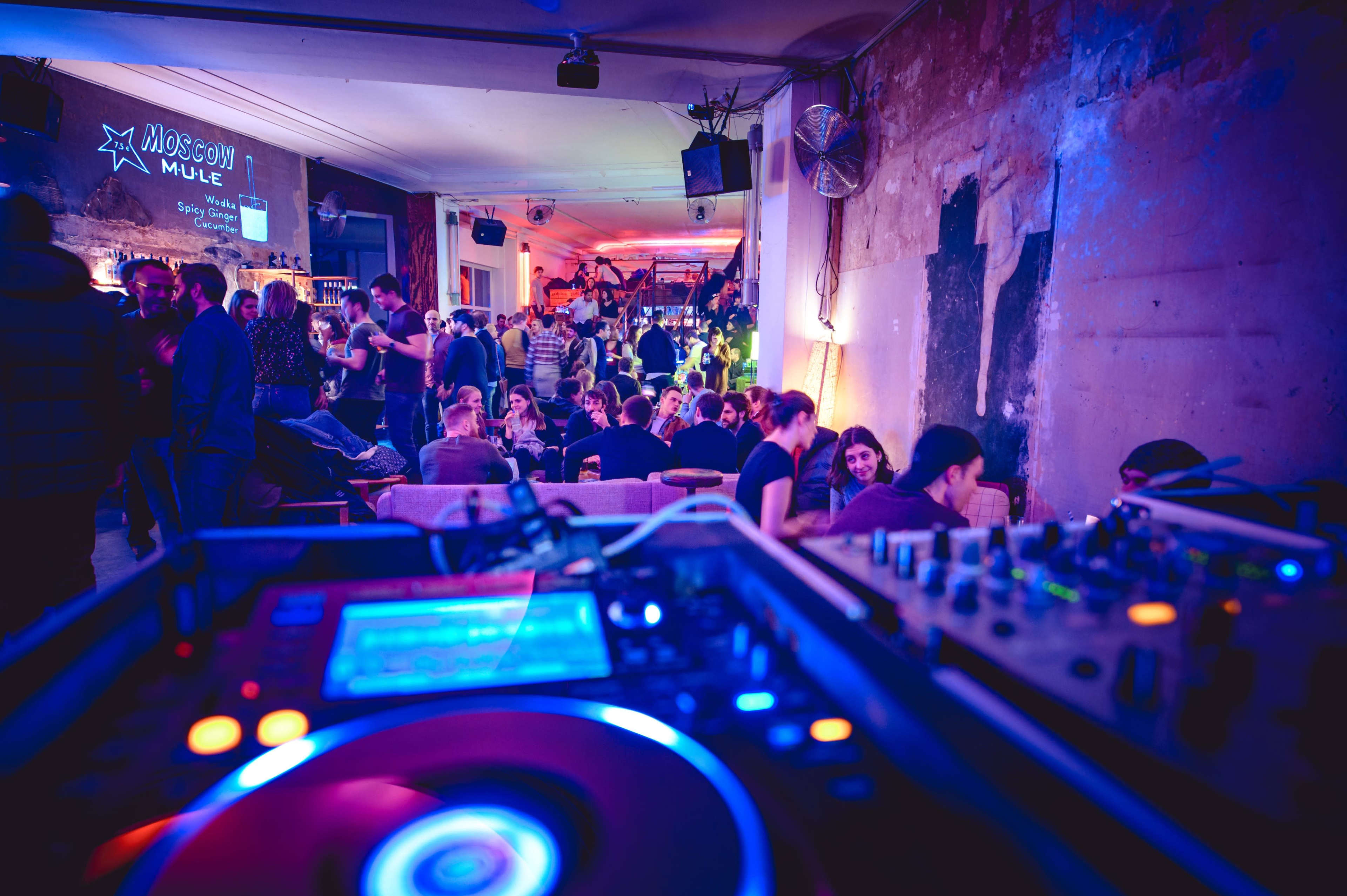 A DJ setup is in the foreground while a crowd enjoys drinks and music in a lively bar setting.