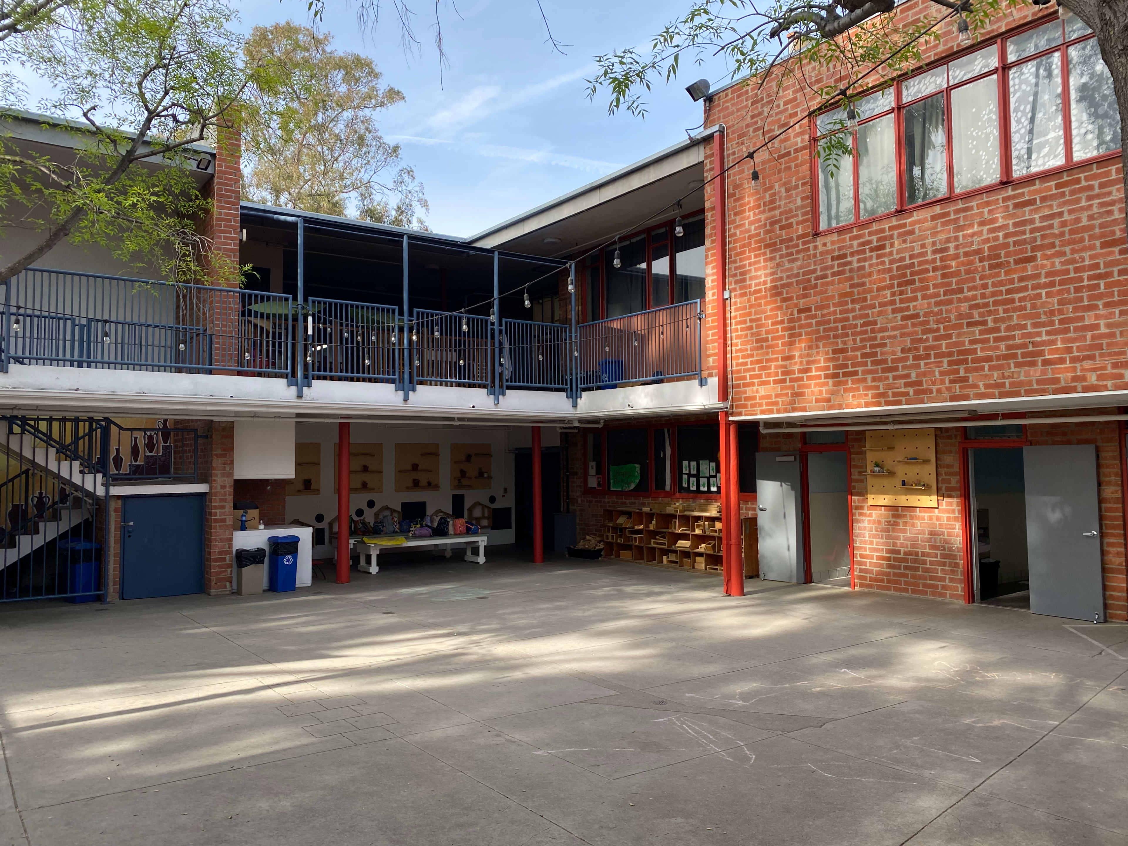 The image shows a school courtyard with two buildings, a table in the center, and seating arranged along one wall.