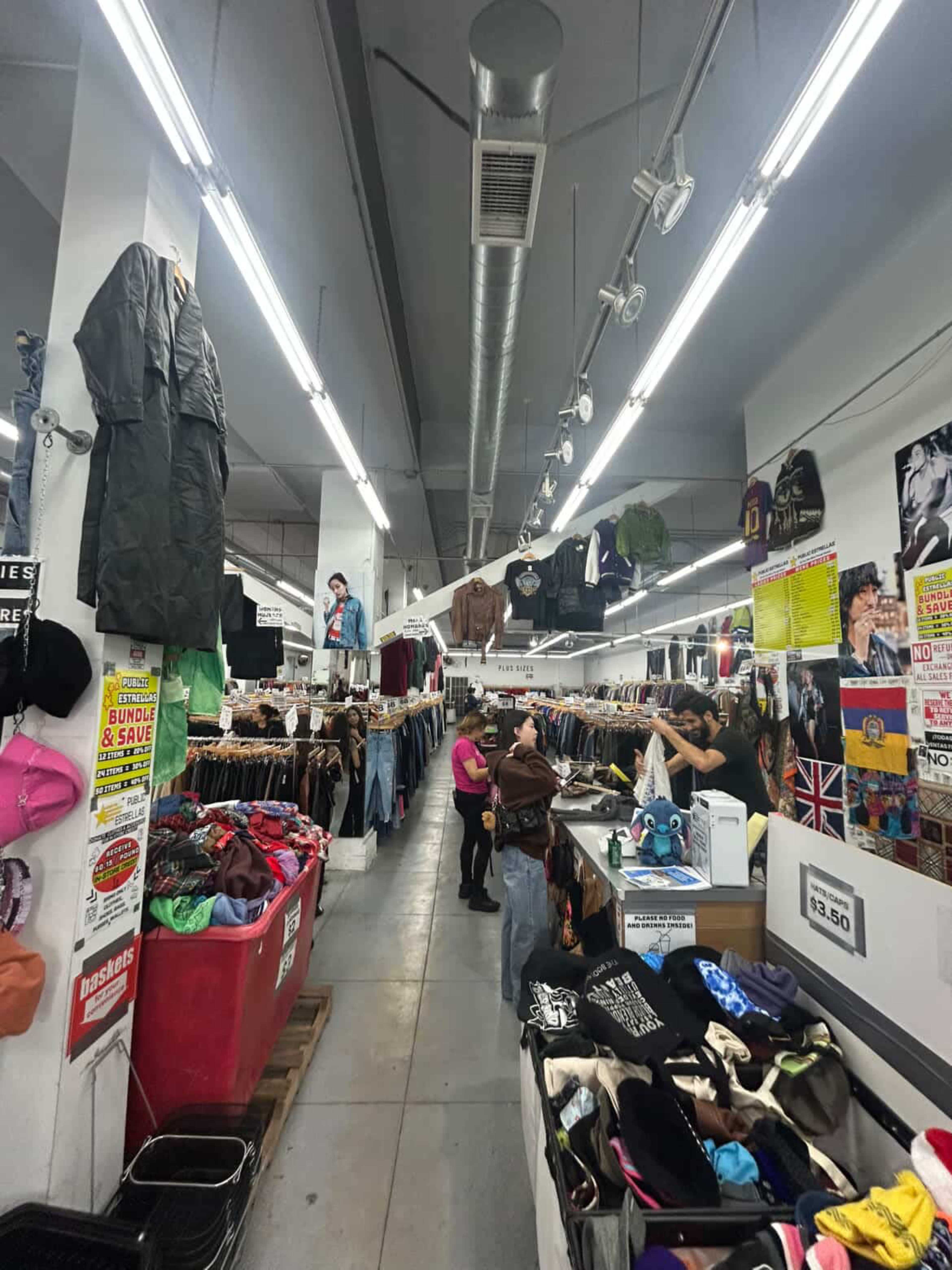 The image shows a bustling thrift store aisle filled with clothing racks, overhead garments, and shoppers browsing among various items.