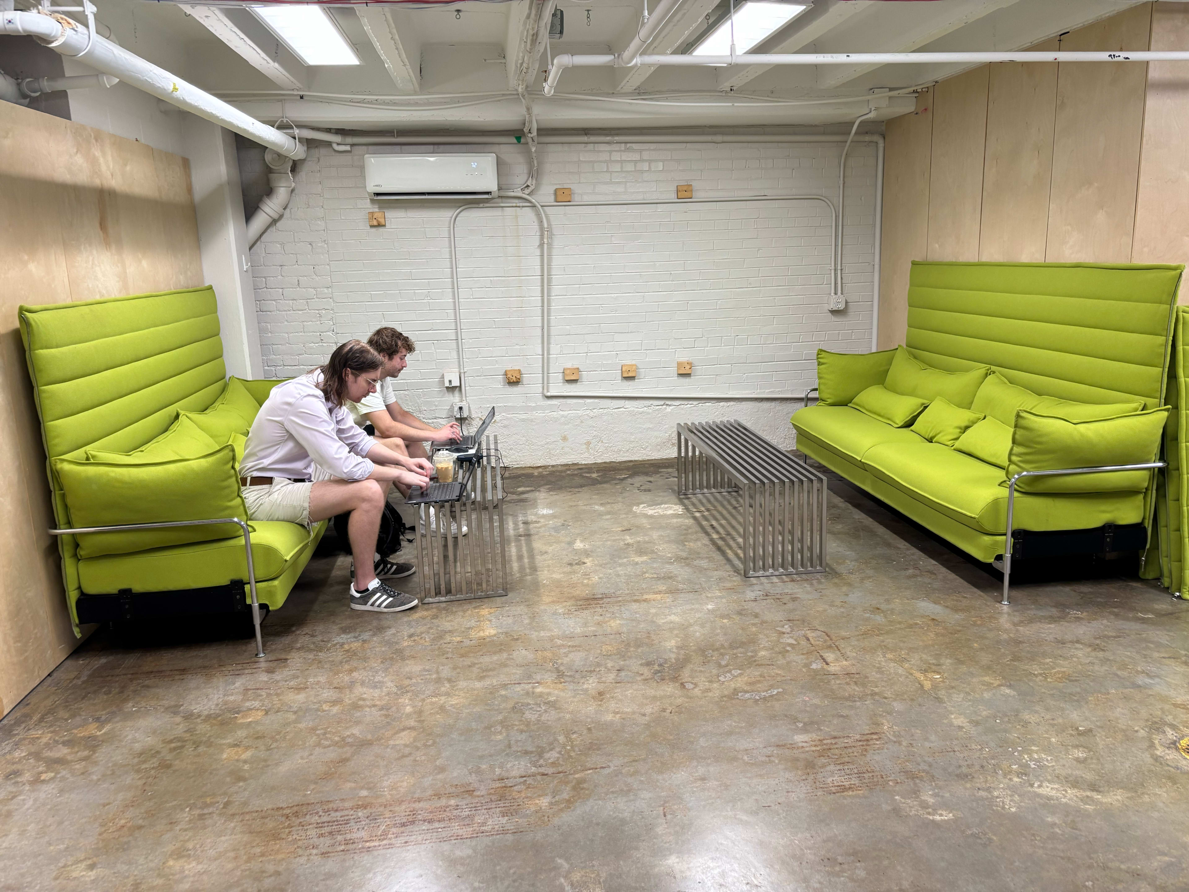 Two individuals are seated on bright green sofas working on laptops at a table in a minimalist, industrial-style room.