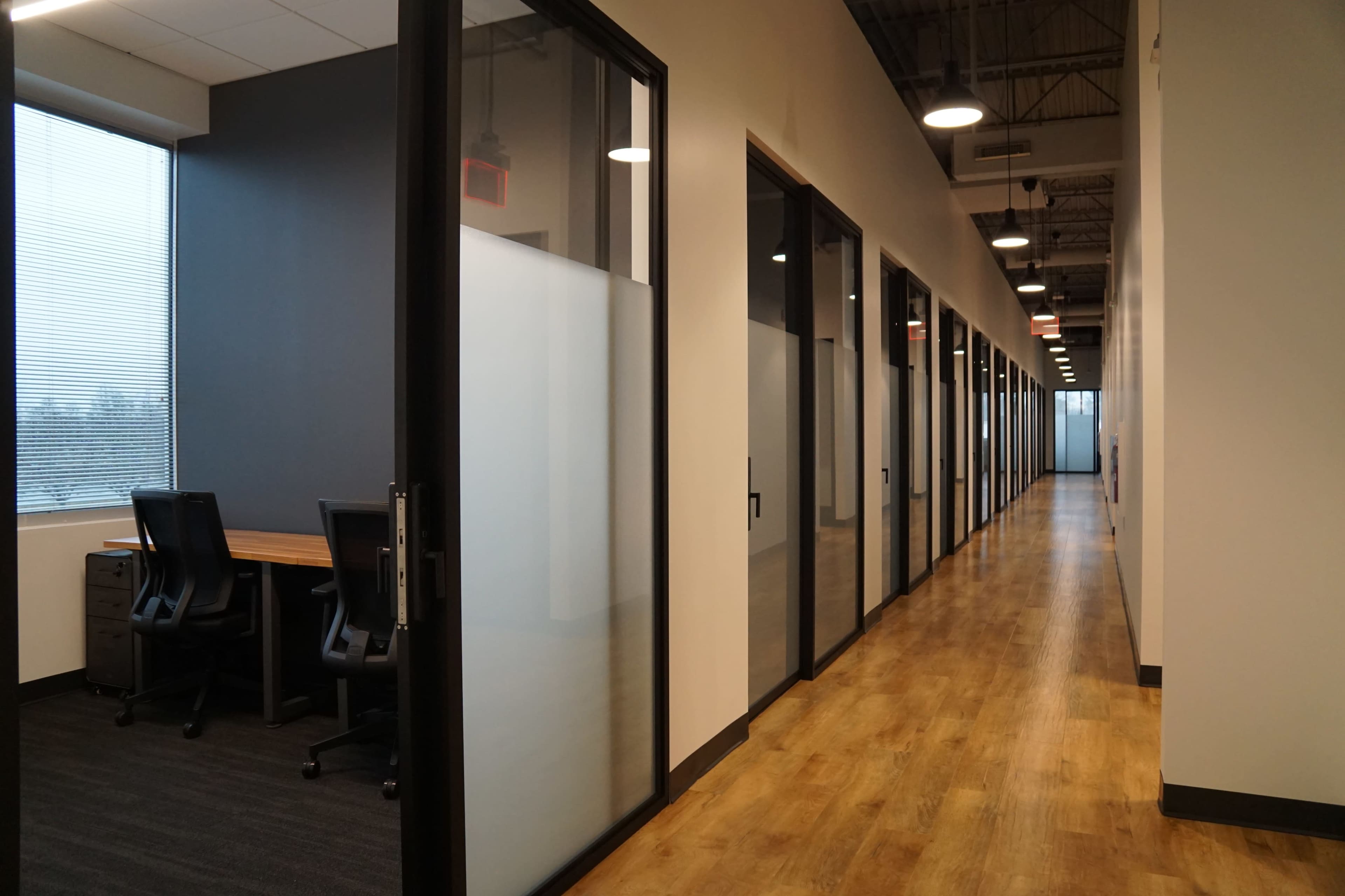 The image shows a modern office hallway with glass-partitioned rooms on either side and wooden flooring.