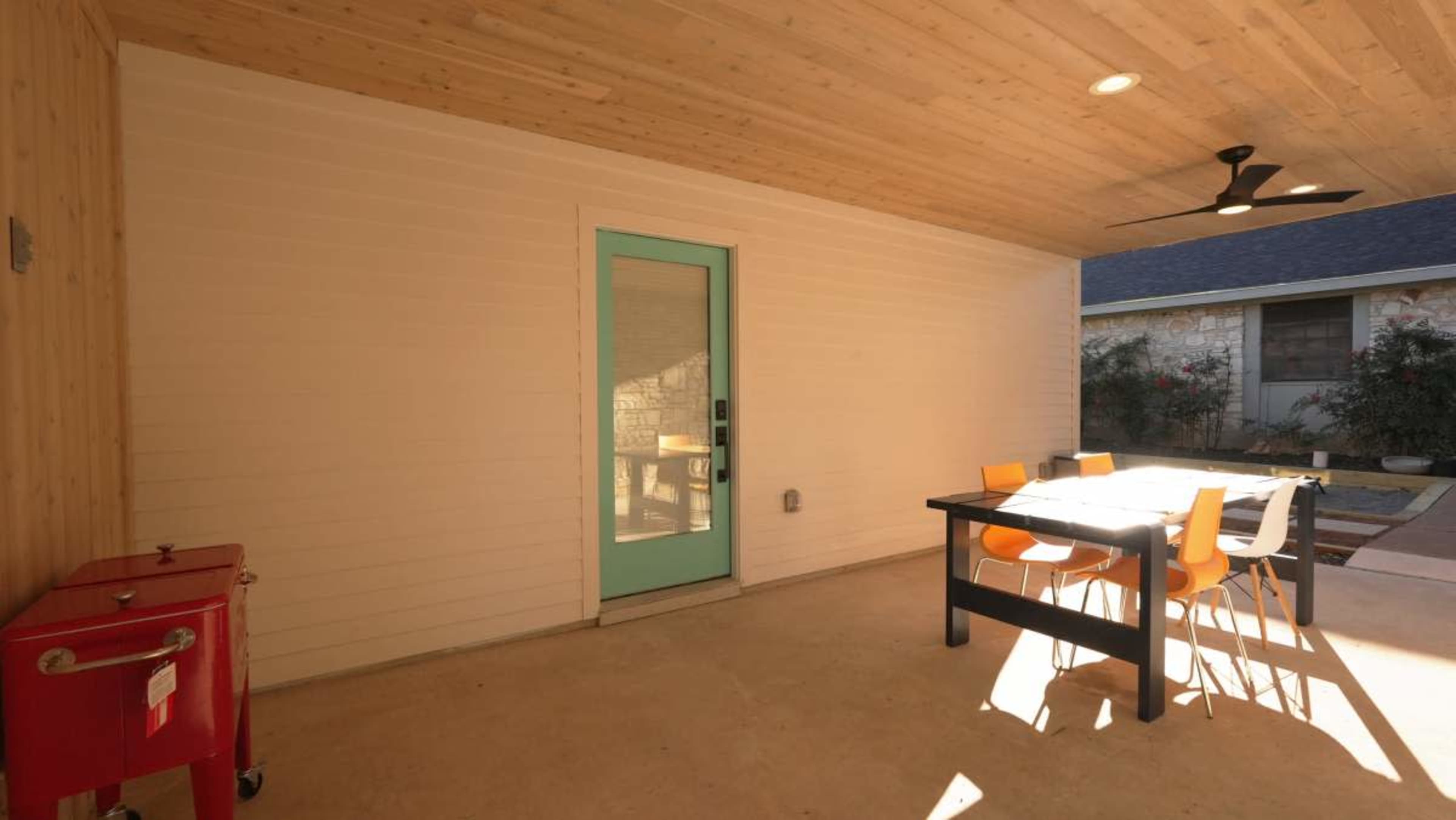 The image shows a covered patio area with a table and chairs, a door with a light teal frame, and a red cabinet against the wall.