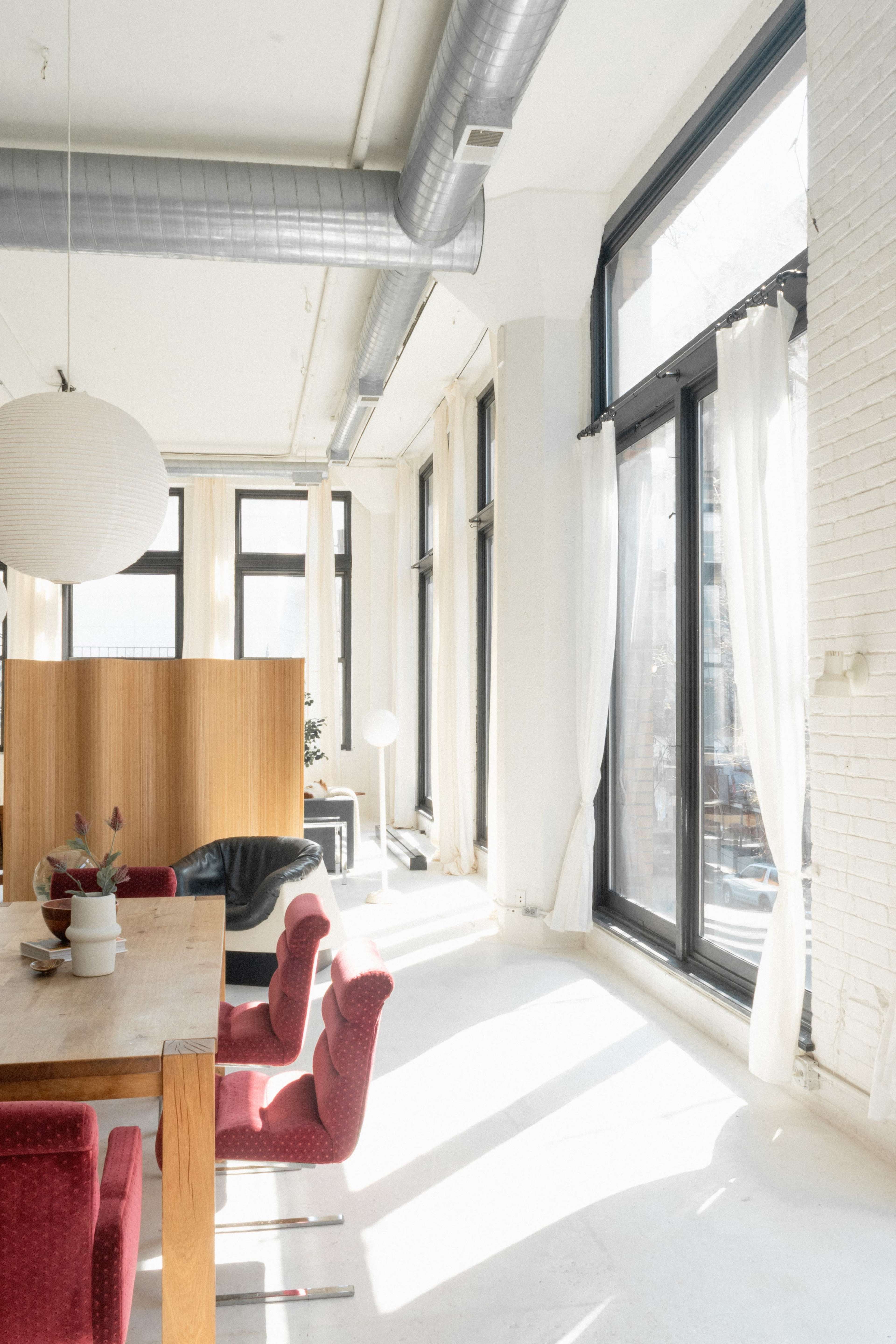 A bright and airy room features a large wooden table with red upholstered chairs and expansive windows allowing natural light to fill the space.