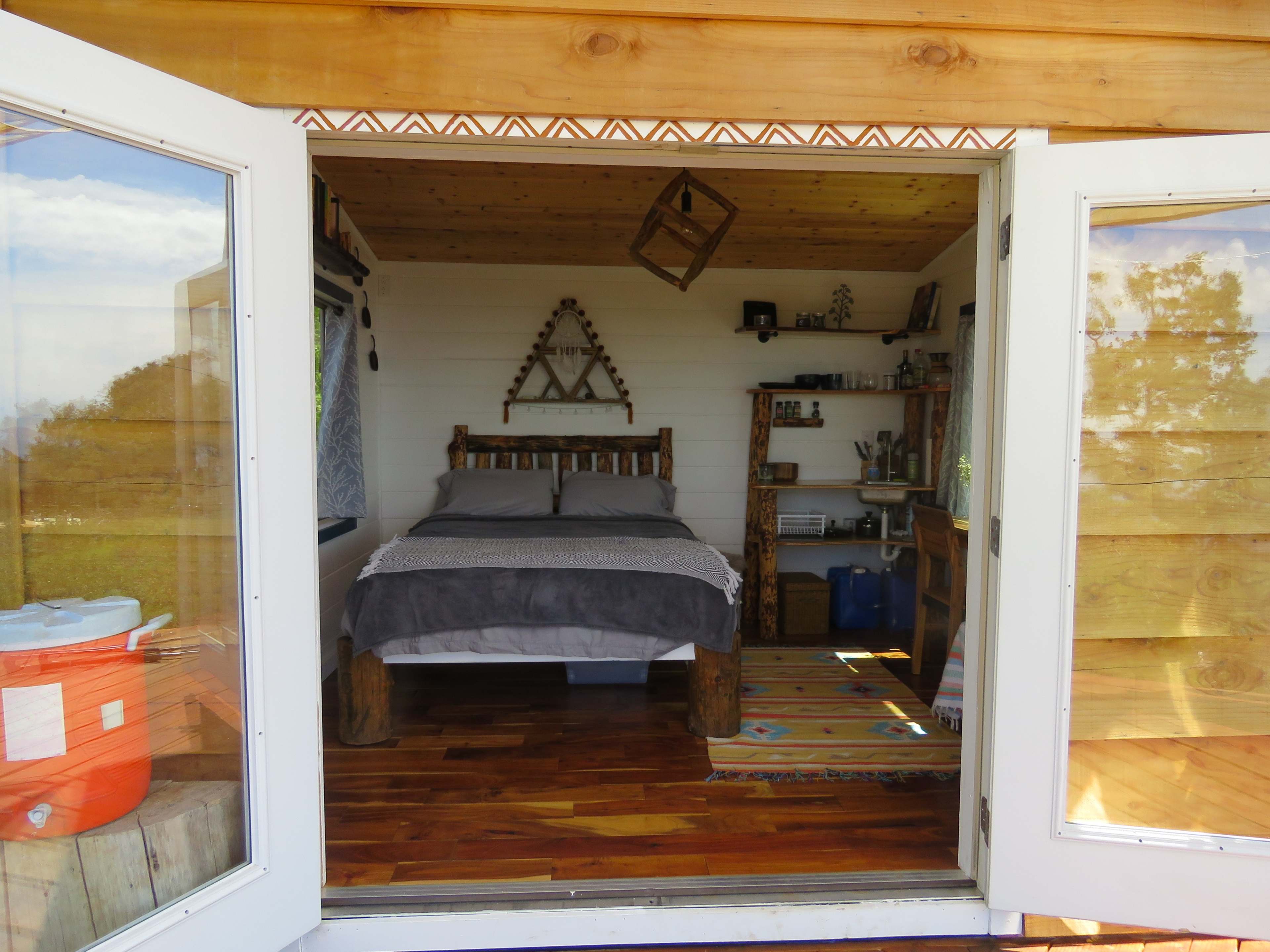 The interior of a small wooden bedroom with a bed, bookshelf, and decorative items framed by open double doors.