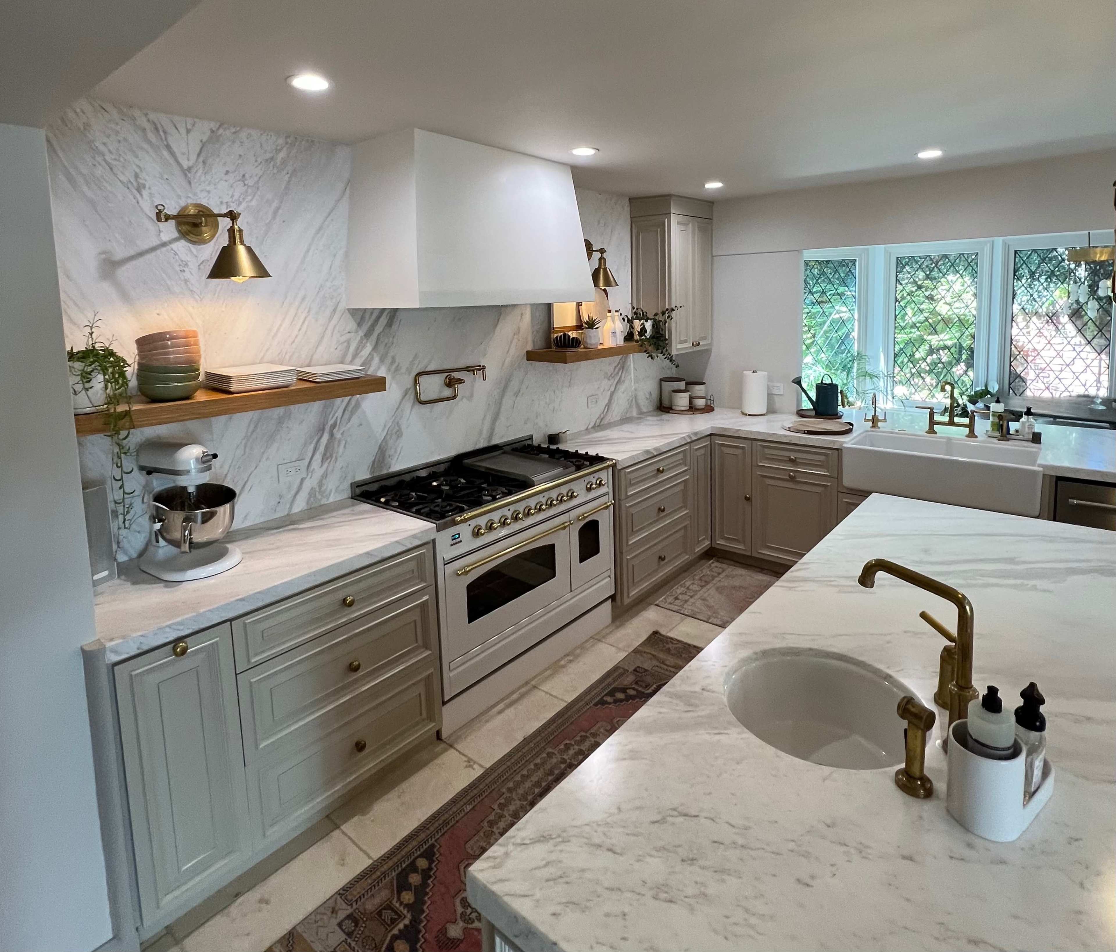 A modern kitchen features marble countertops, white cabinetry, and stainless steel appliances, with large windows providing natural light.