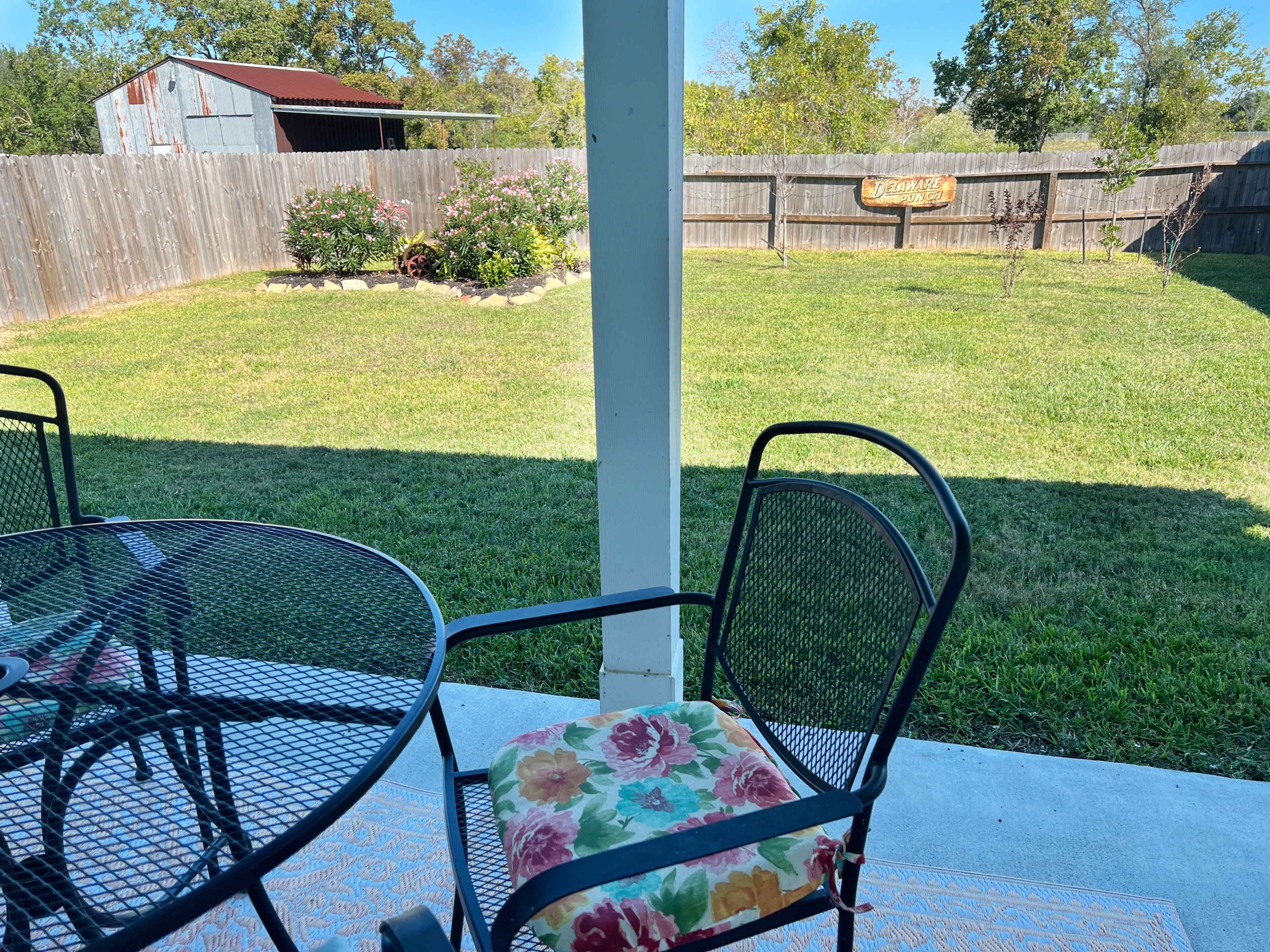 A patio area with a black metal table and chair is set up in front of a grassy backyard featuring a wooden fence and a small garden.