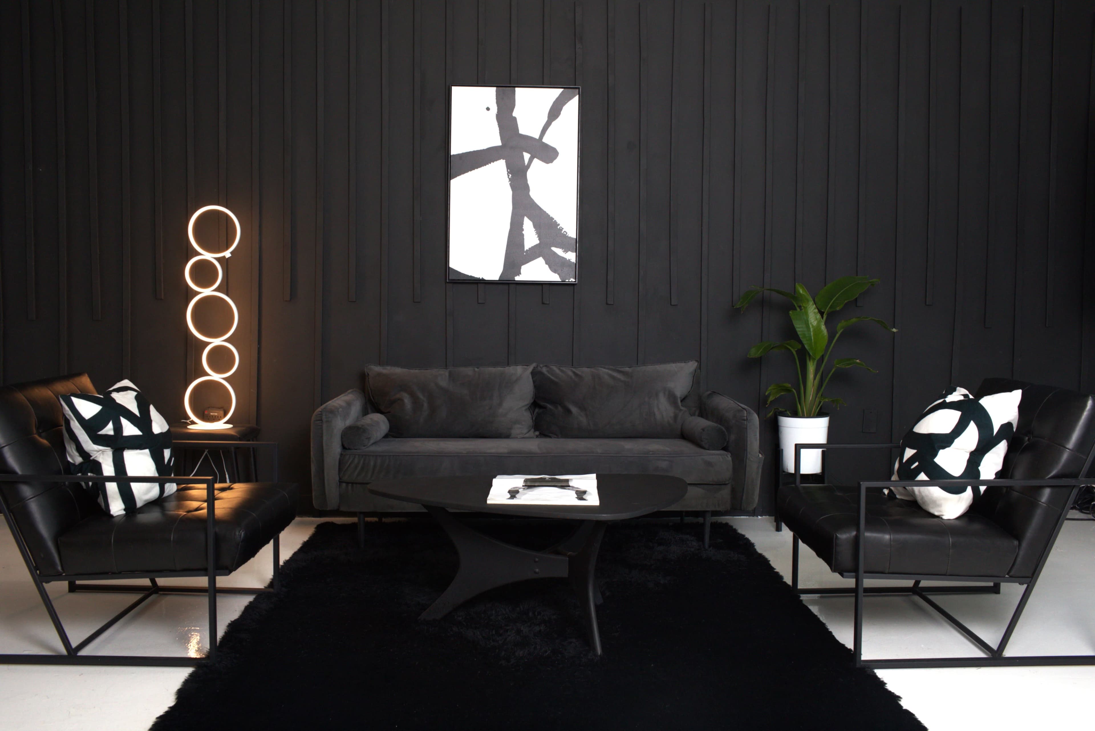 A modern living room with a black sectional sofa, two black leather chairs, a circular coffee table, and a plant, all set against a black-paneled wall and a piece of abstract wall art.