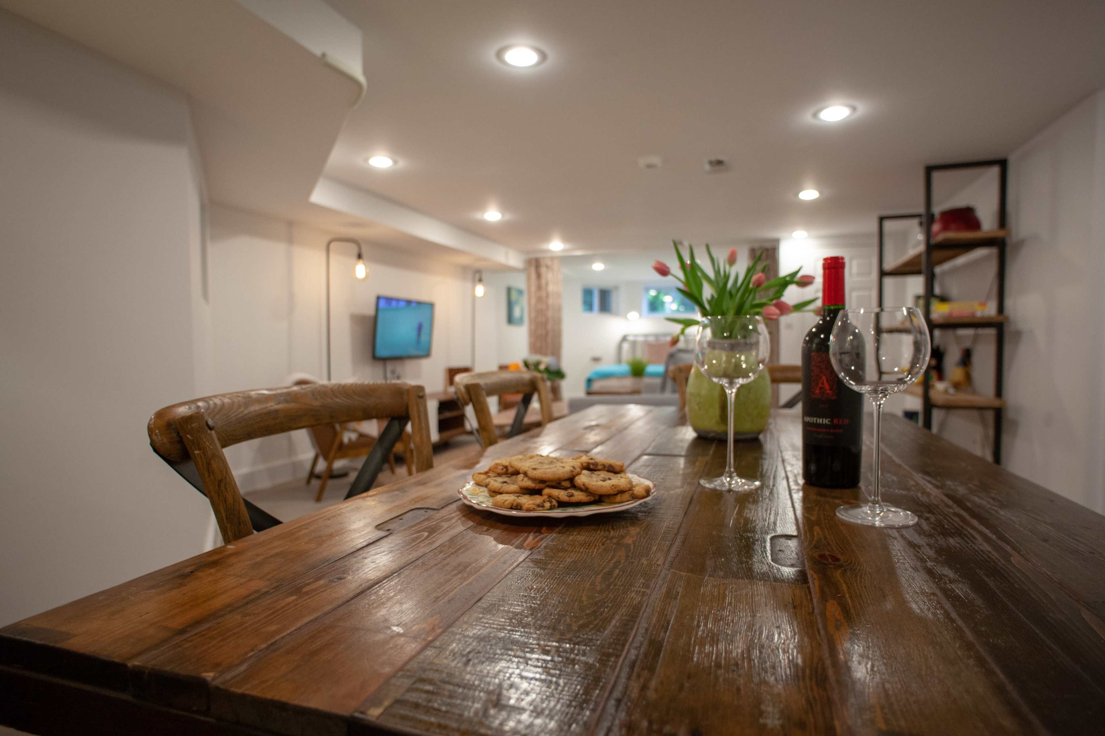 A wooden dining table is set with a plate of cookies, a bottle of wine, and two glasses, while a cozy living area is visible in the background.