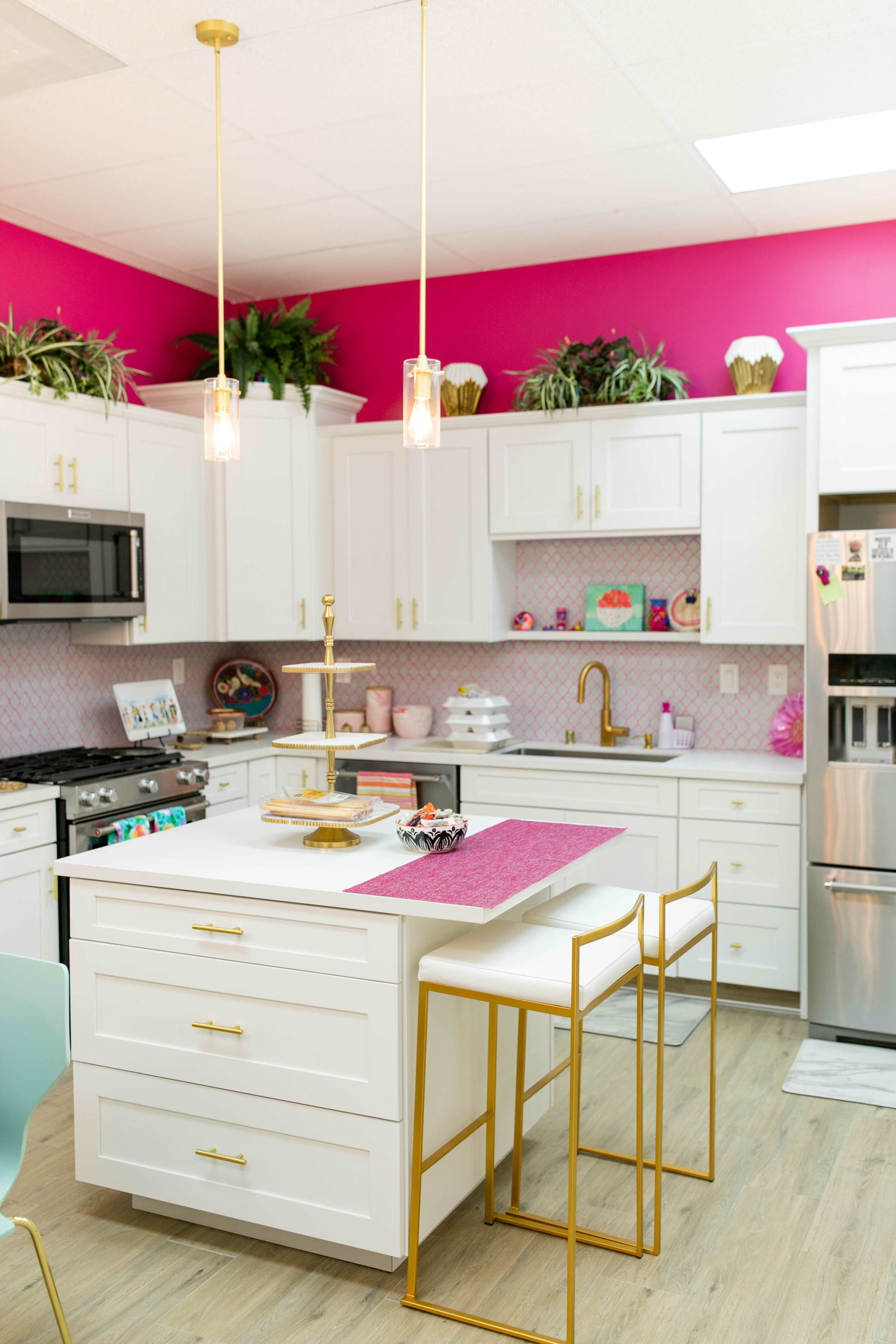 The kitchen features white cabinetry, gold accents, and bright pink walls, along with a central island and modern appliances.