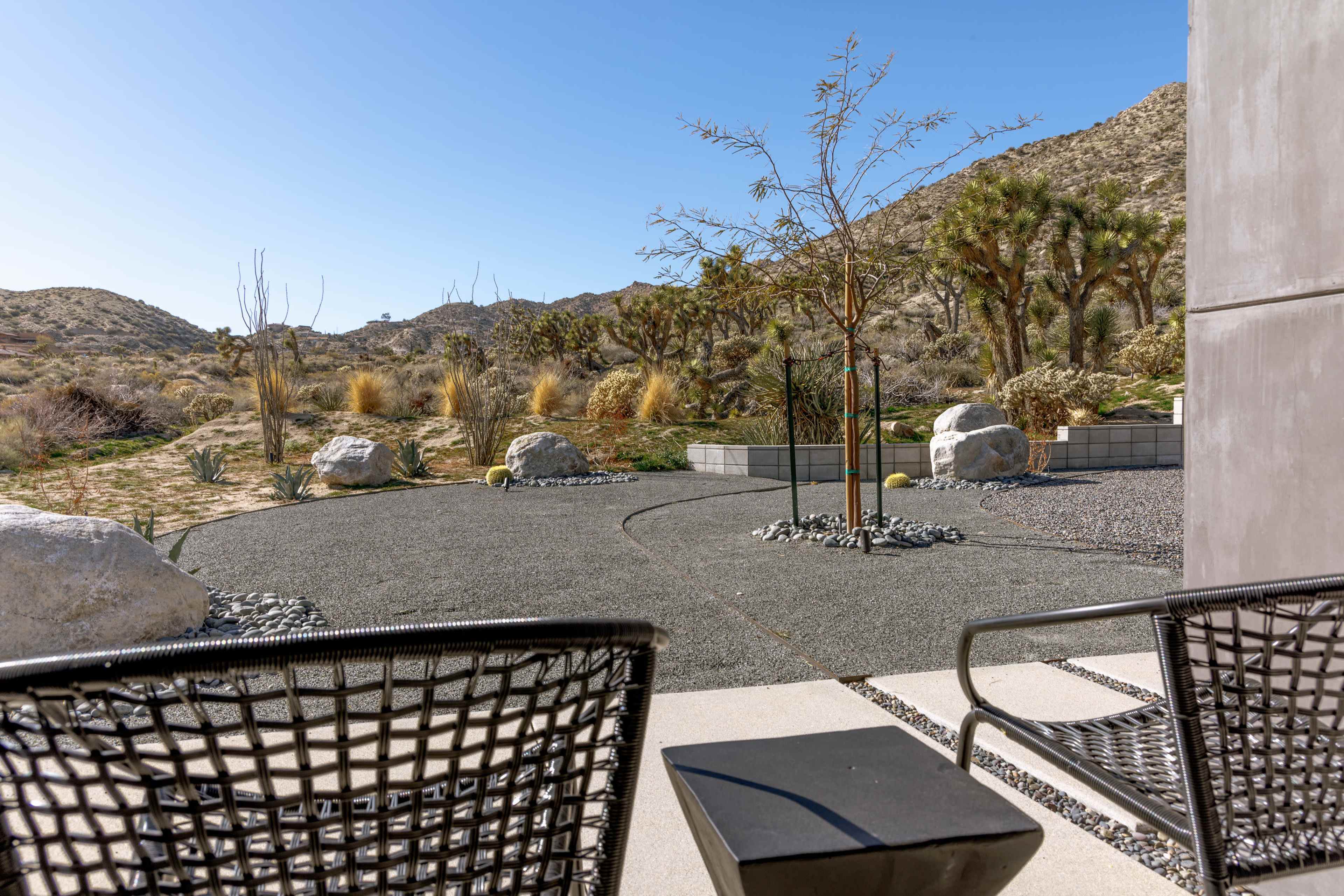 The image shows a desert landscape view from a patio featuring black chairs, gravel pathways, and various plants and rocks.