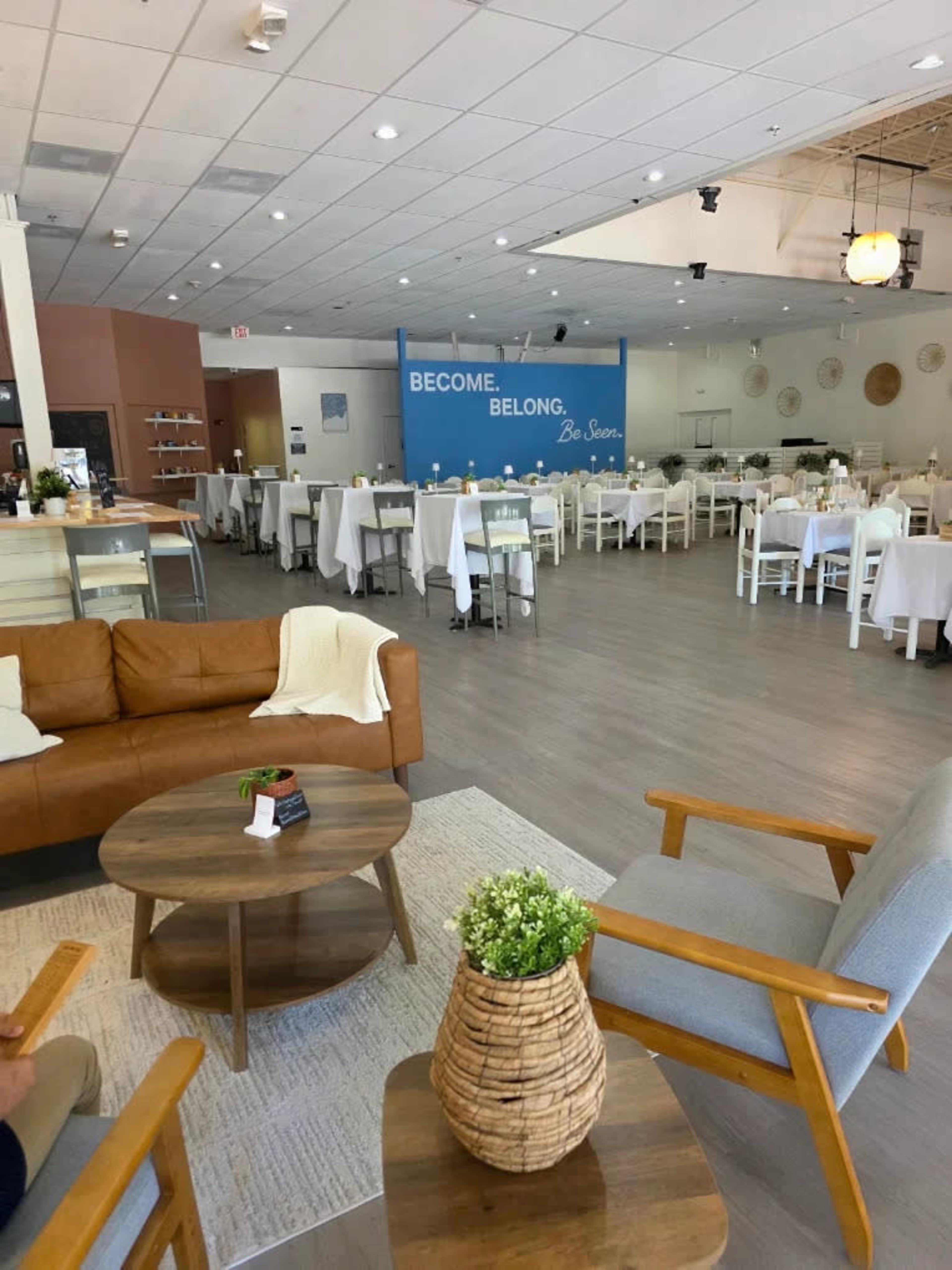 The image shows a modern, spacious restaurant interior with white tablecloths, wooden furniture, and a wall that has the text "BECOME. BELONG. Be Seen." prominently displayed.