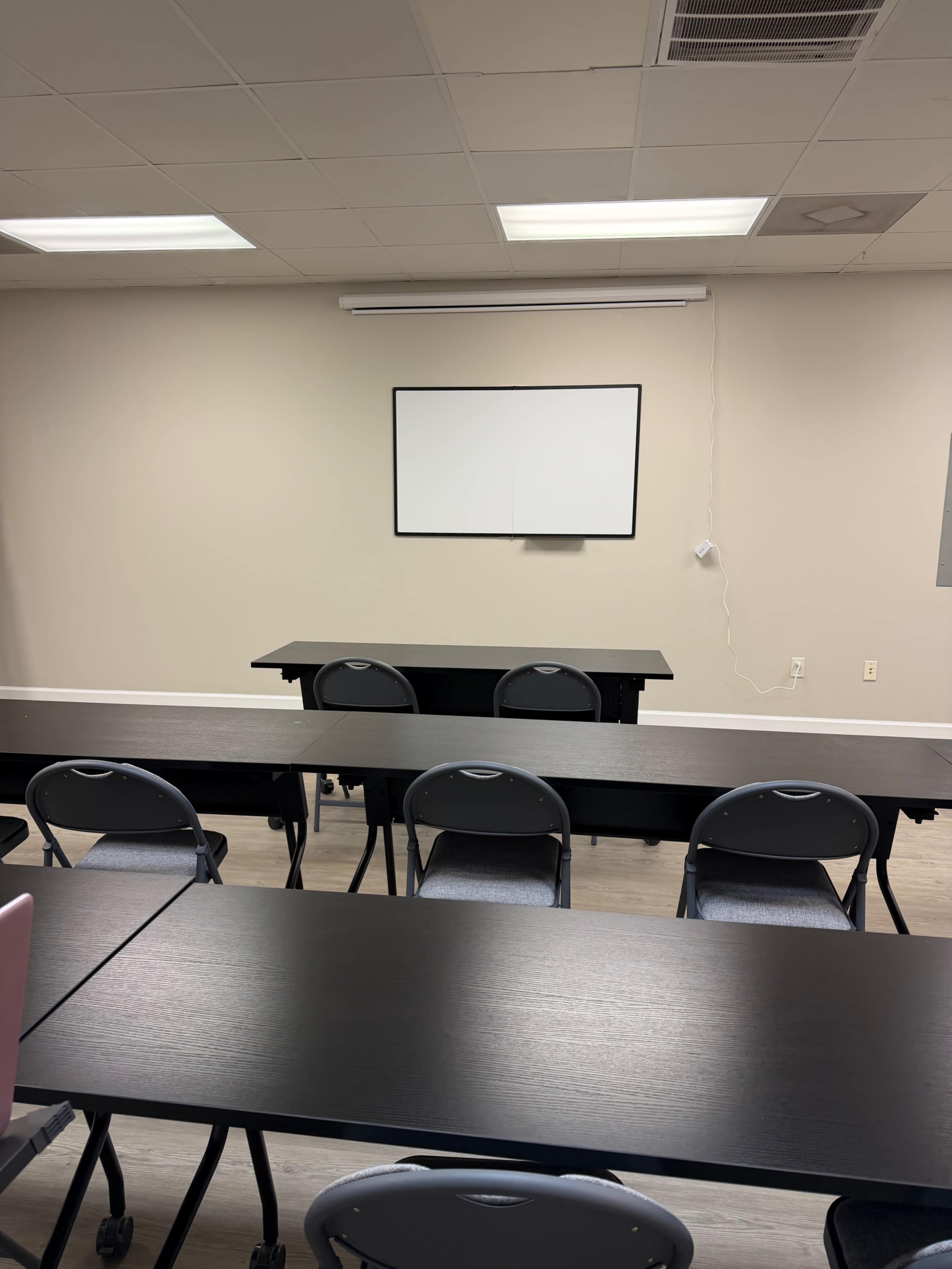 The image shows a classroom with rows of black tables and chairs arranged in front of a blank whiteboard on the wall.