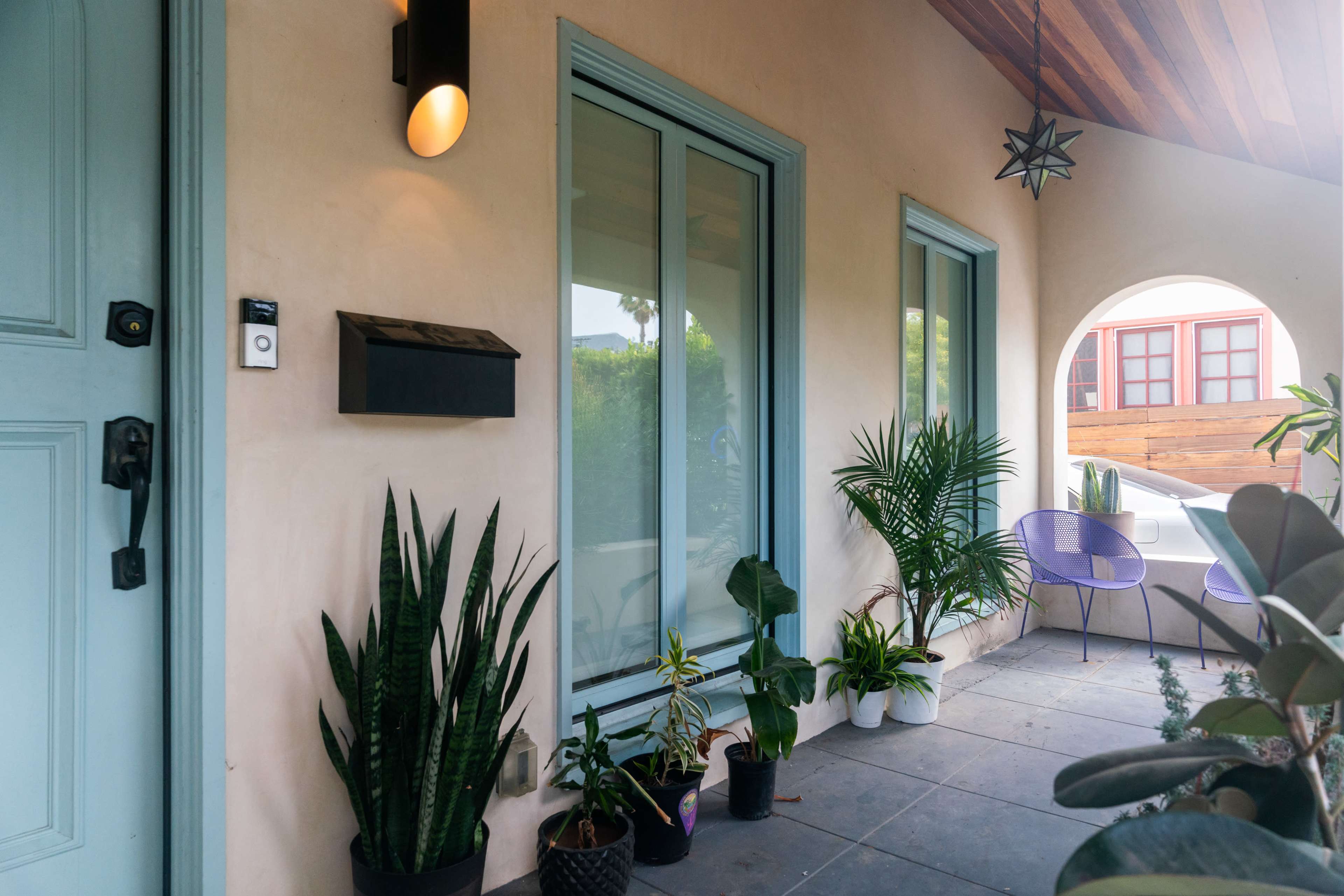 The image shows a home entrance with a light blue door, potted plants along the side, and two purple chairs positioned on a covered porch.