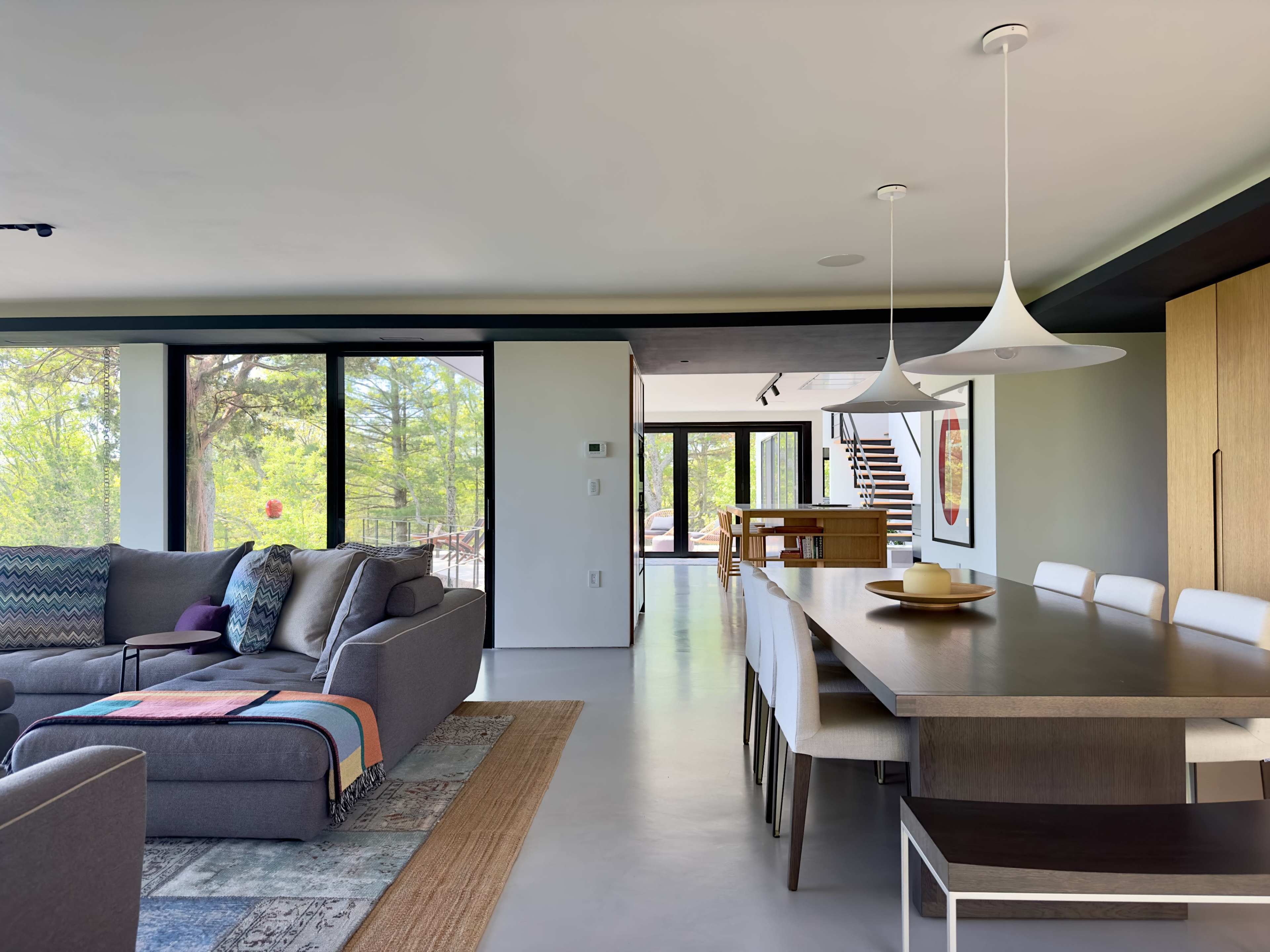 The image shows a modern living space featuring a gray sofa, a long dining table, and large windows with a view of greenery outside.