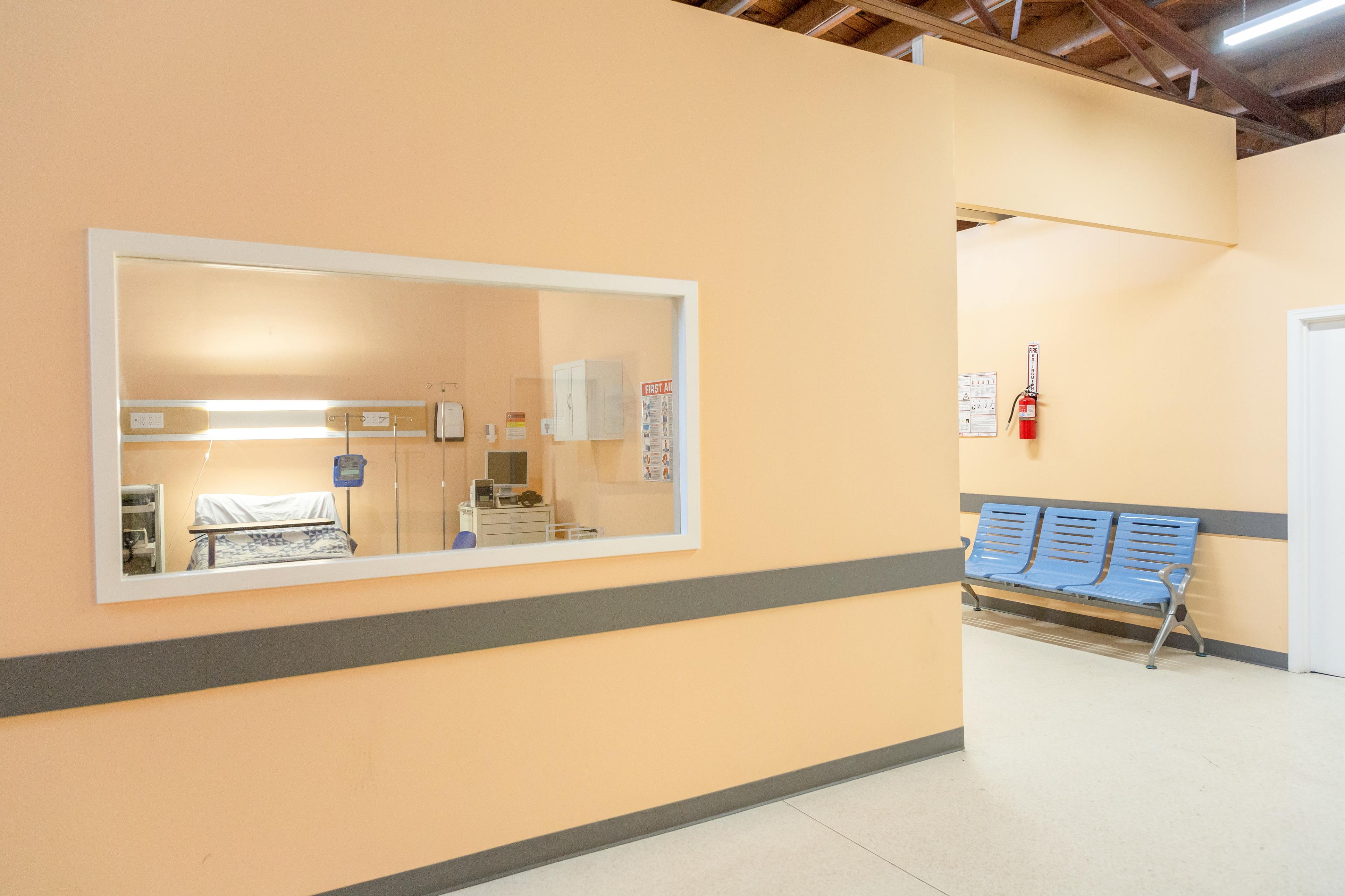 A hospital corridor with a window looking into a patient room, a seating area, and orange-colored walls.