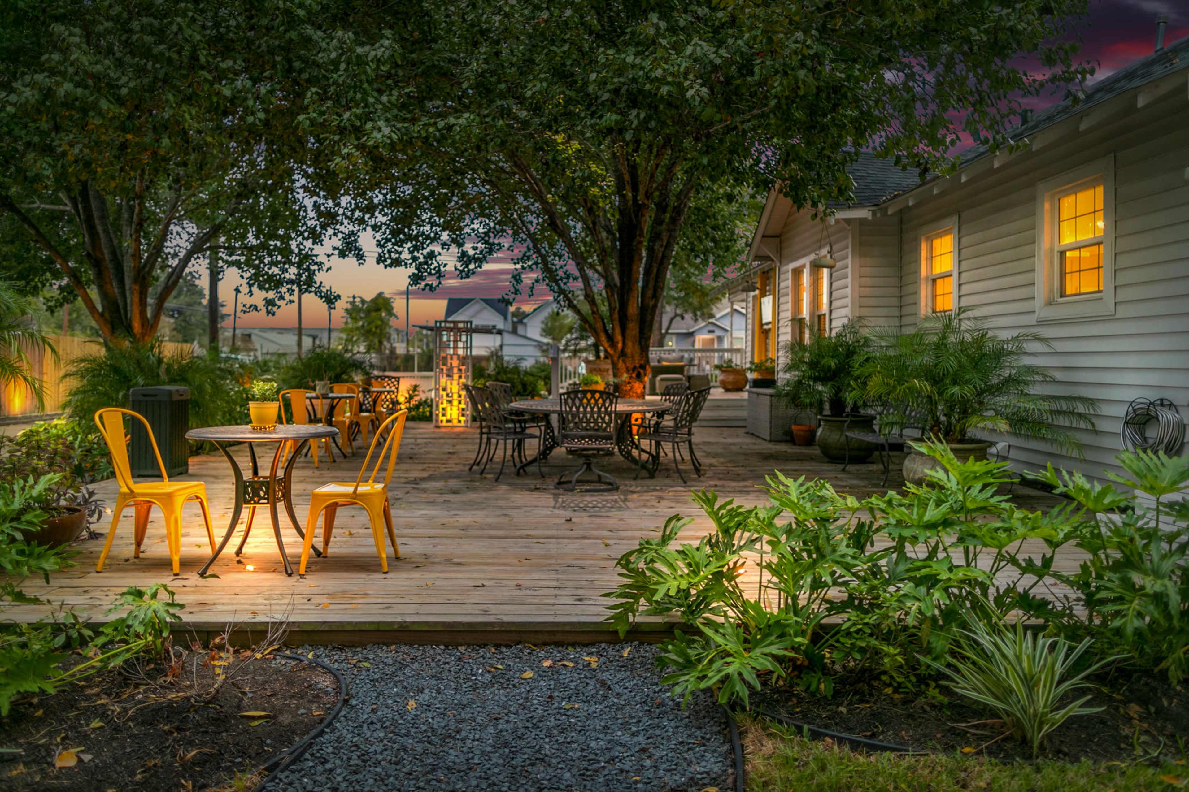 A wooden deck with several tables and chairs surrounded by plants and trees, illuminated by warm lighting from the windows of a nearby house.