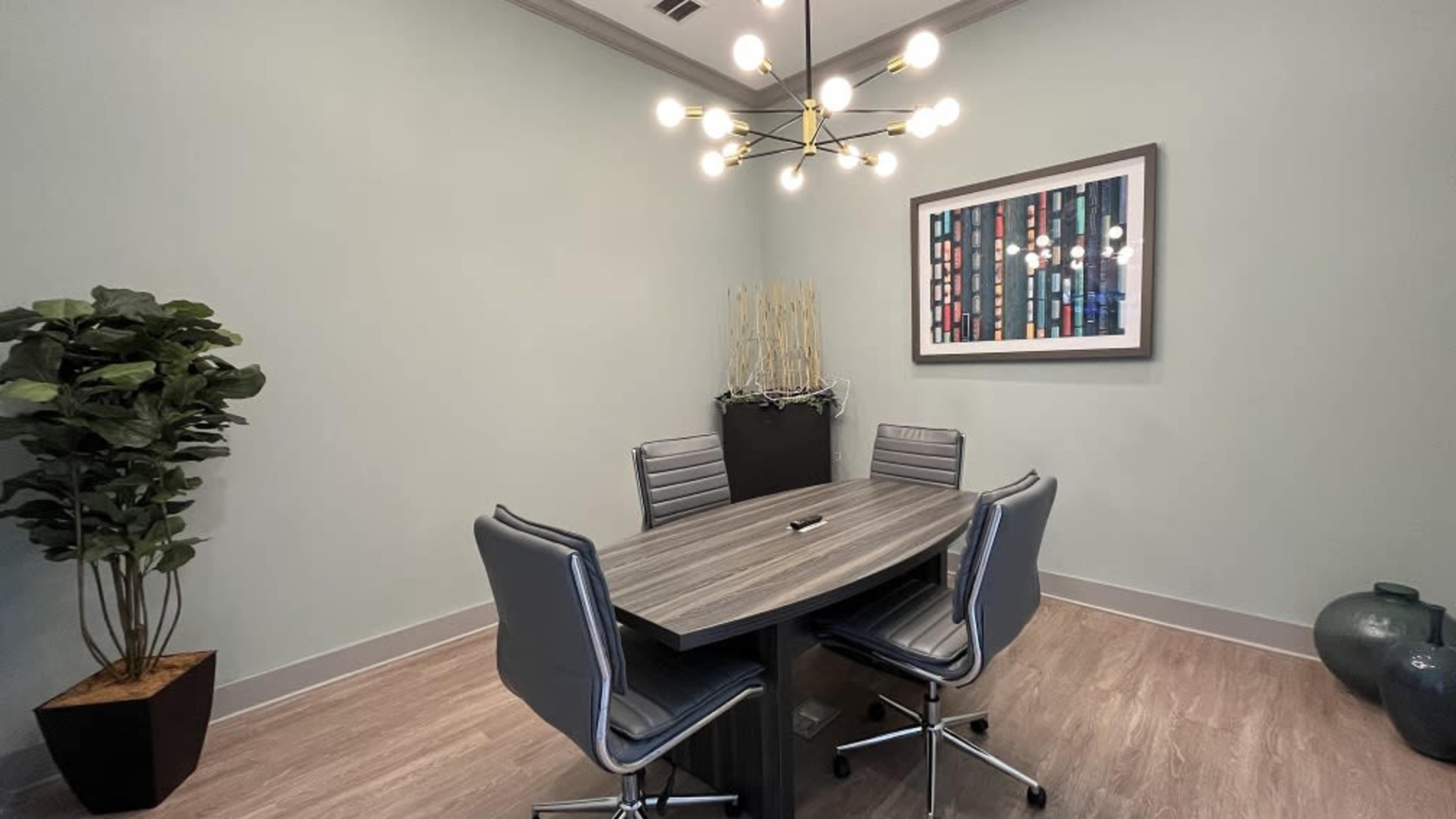 A small conference room with a modern oval table surrounded by four steel-framed chairs, a decorative plant, and a colorful artwork on the wall.