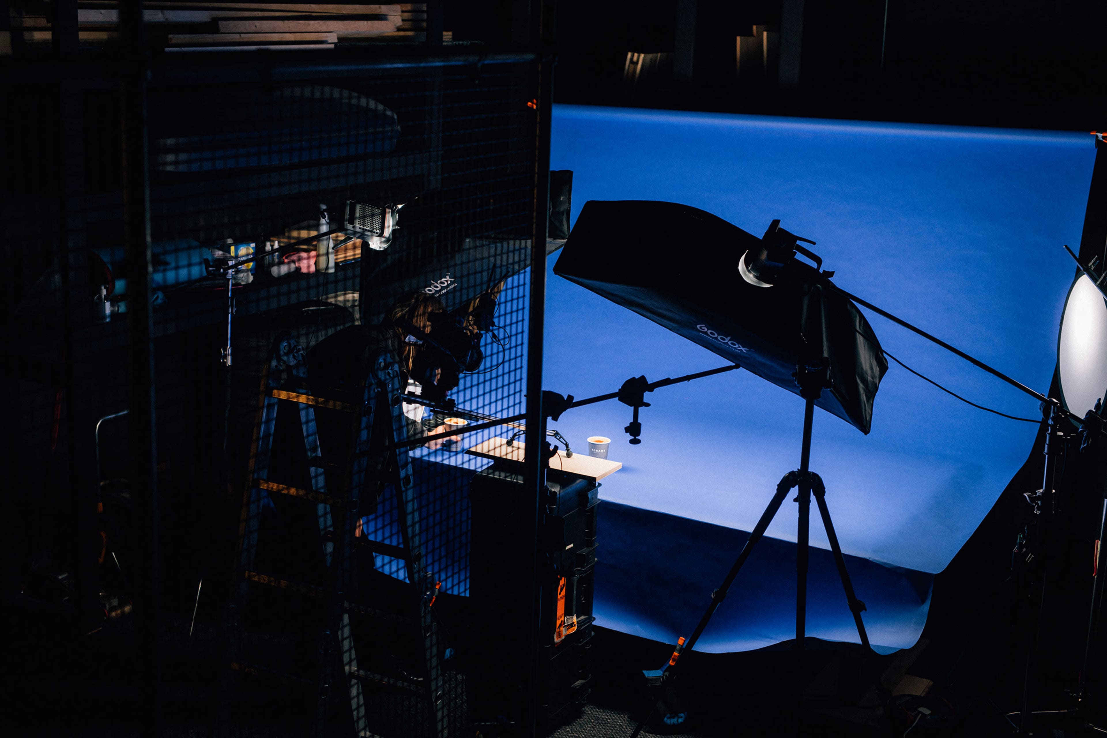 A photographer is positioned beside a ladder, adjusting equipment in a studio with a blue backdrop and various lighting setups.
