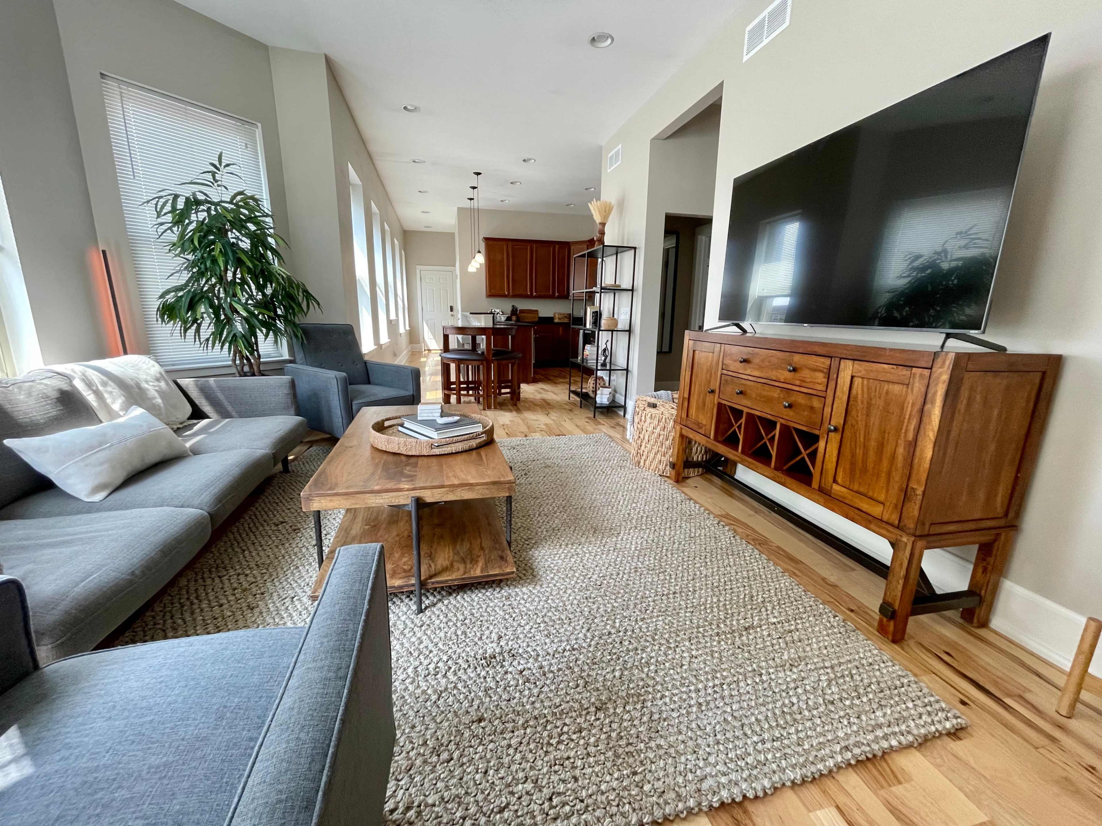 A modern living room features a gray sofa, a wooden coffee table, a large television, and a dining area visible in the background.