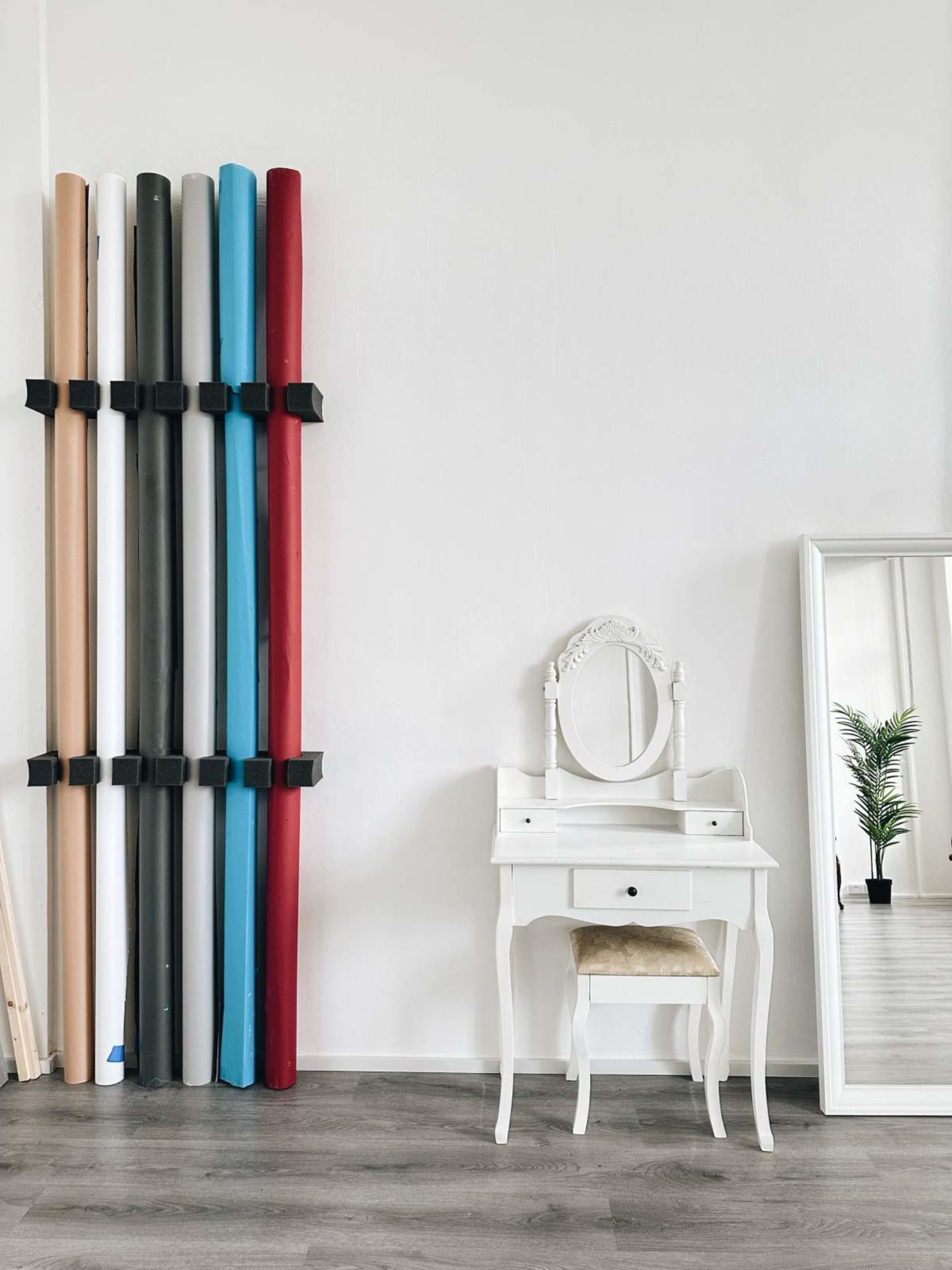 The image features a neatly arranged set of rolled backgrounds in various colors against a white wall, alongside a white vanity table with a round mirror and a small potted plant visible in the reflection.