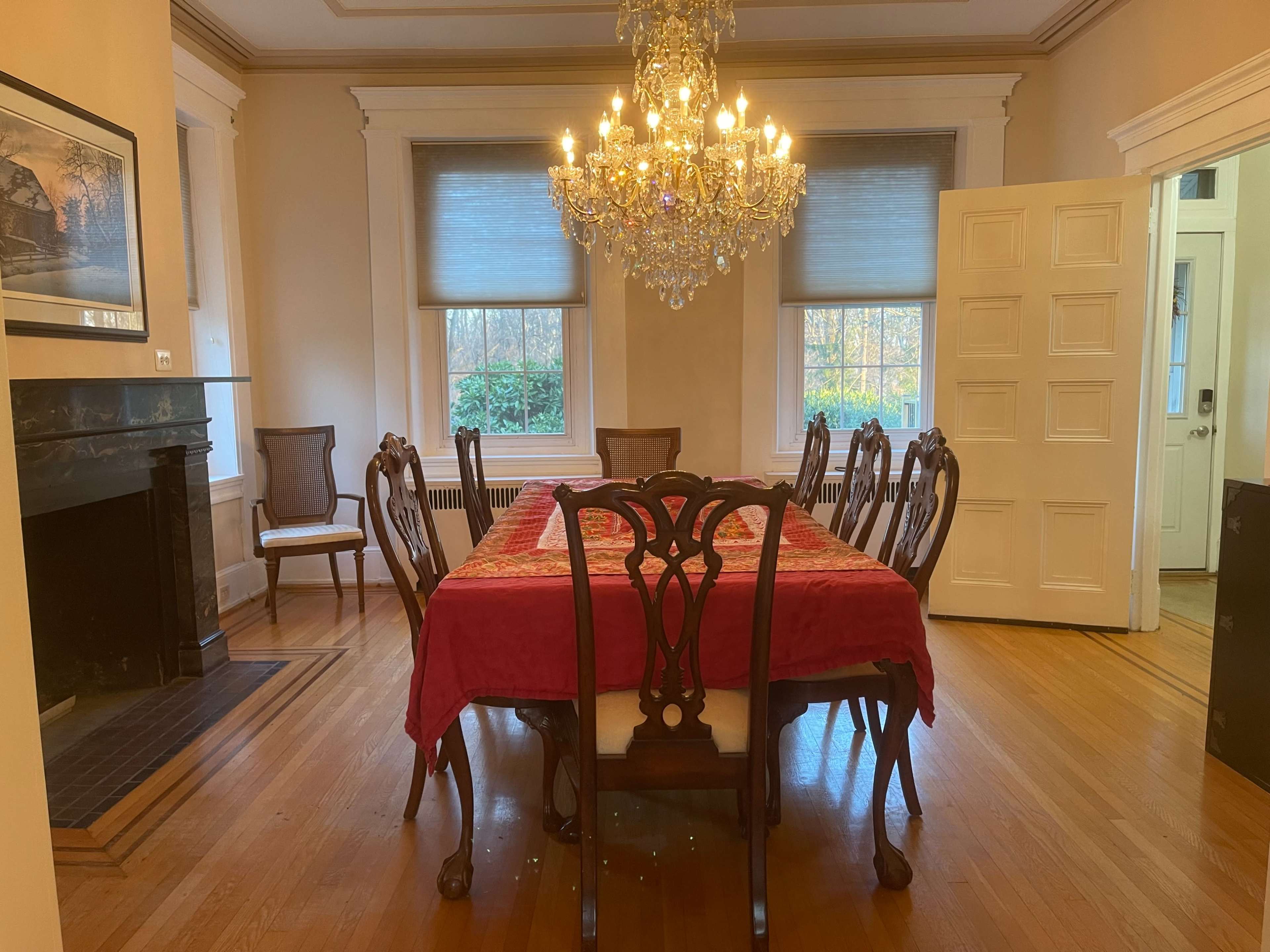 A large dining room features a wooden table surrounded by eight chairs, illuminated by a chandelier, with a fireplace and two windows in the background.