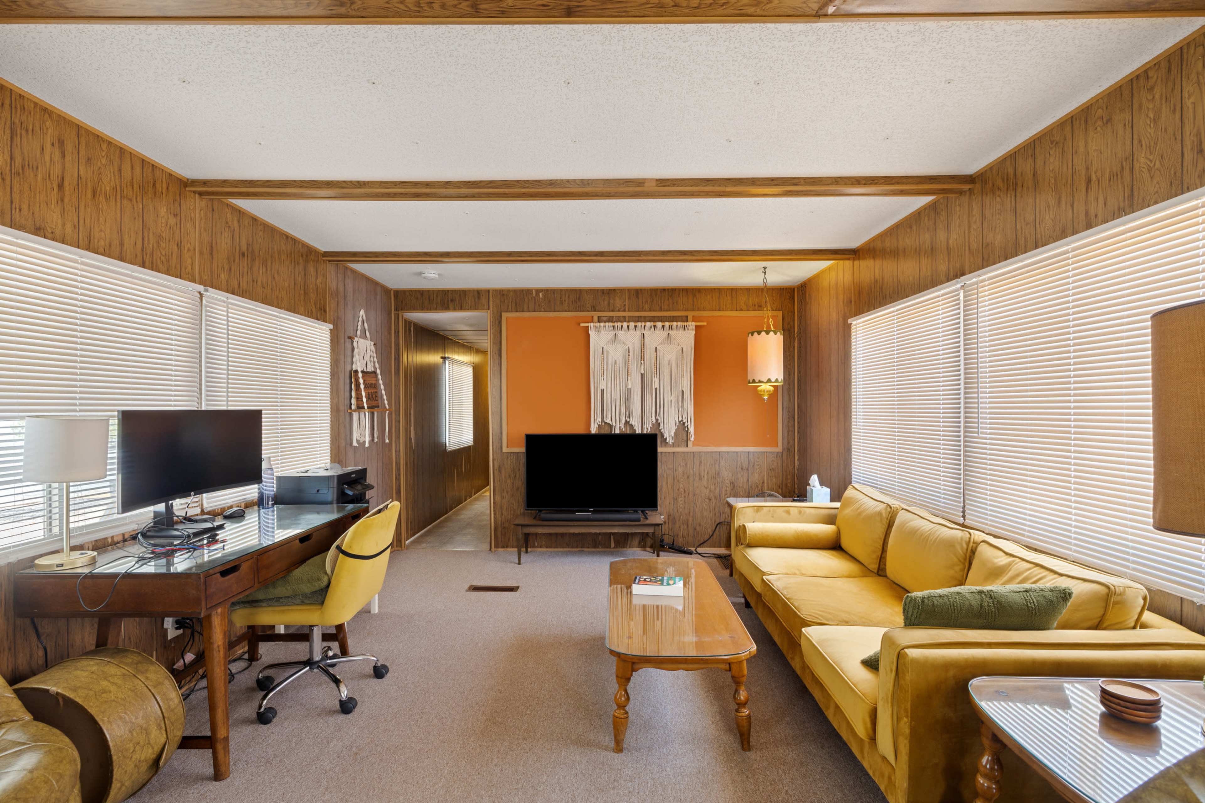 The image shows a cozy living room featuring a yellow sofa, a wooden coffee table, and a desk with a computer across from a television.