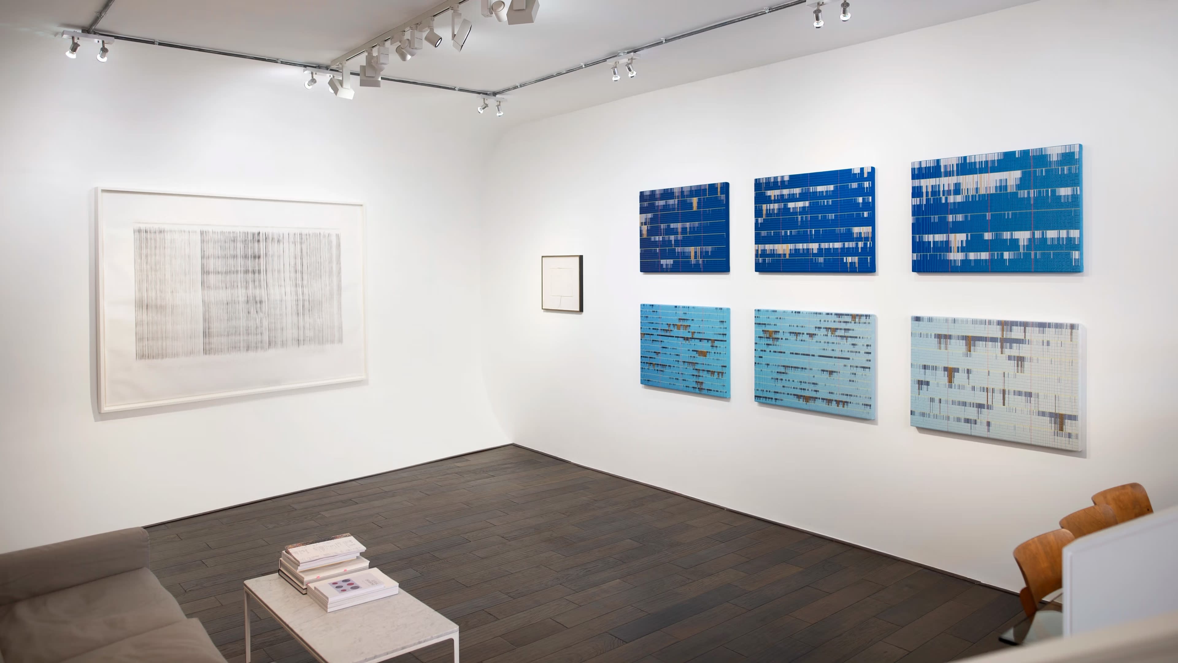 The image shows a modern art gallery featuring a collection of six blue abstract paintings on one wall and a framed artwork on another, with a sofa and a small table in the corner.