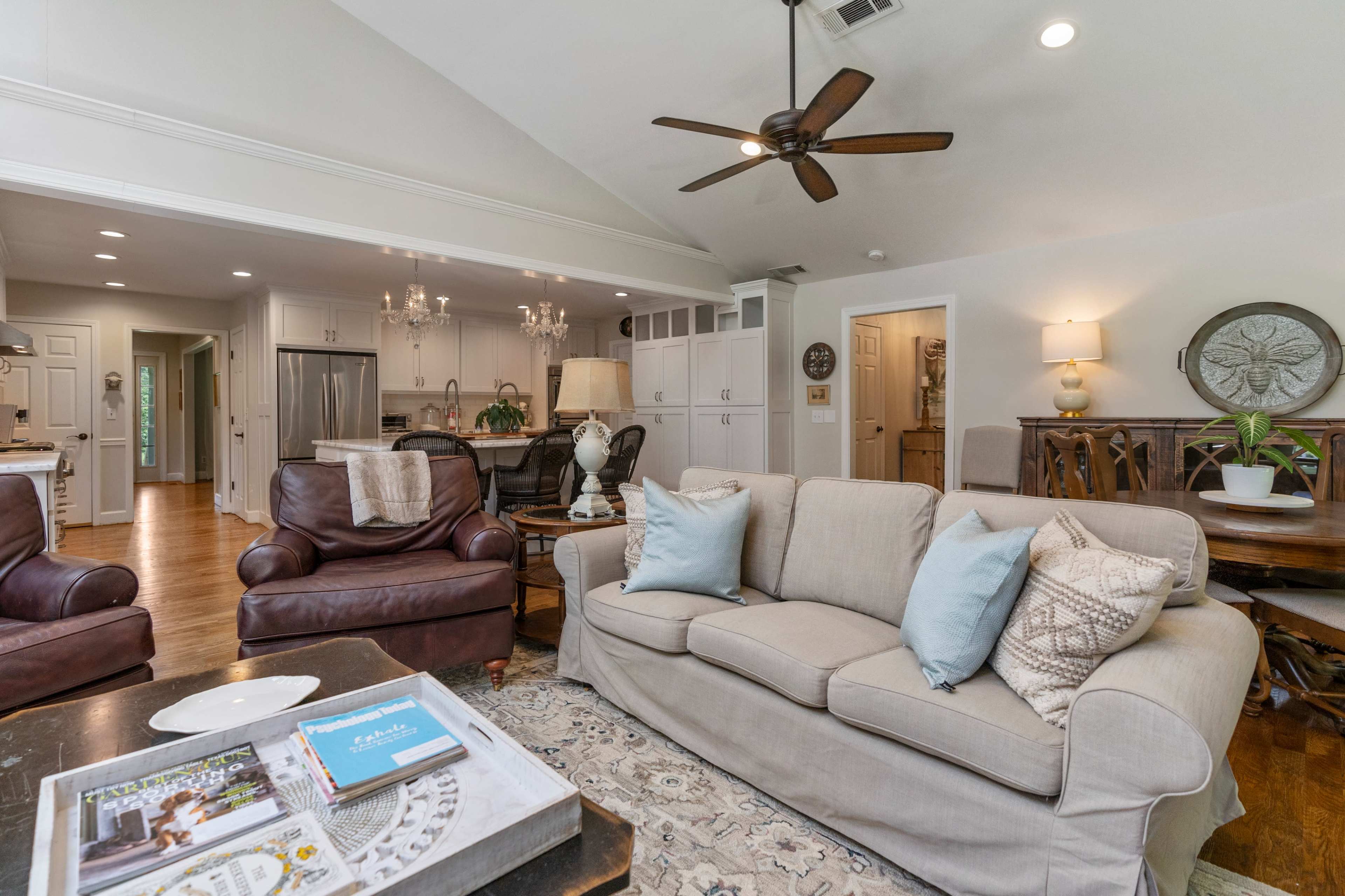A spacious living room with a light-colored sofa, brown leather chairs, and a dining area in the background featuring a wooden table and chandelier.