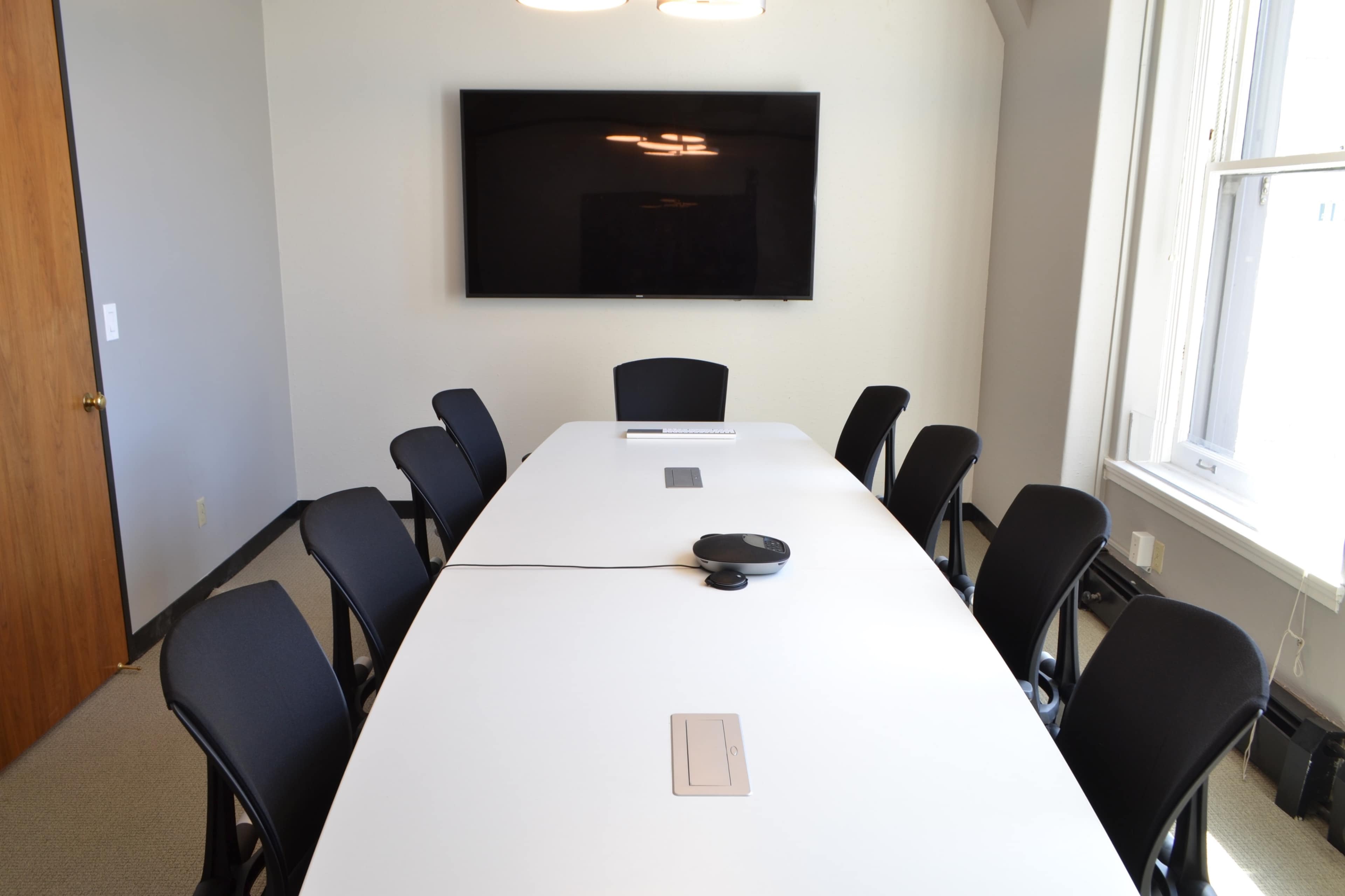 A conference room features a long white table surrounded by eight black chairs and a large flat-screen TV on the wall.
