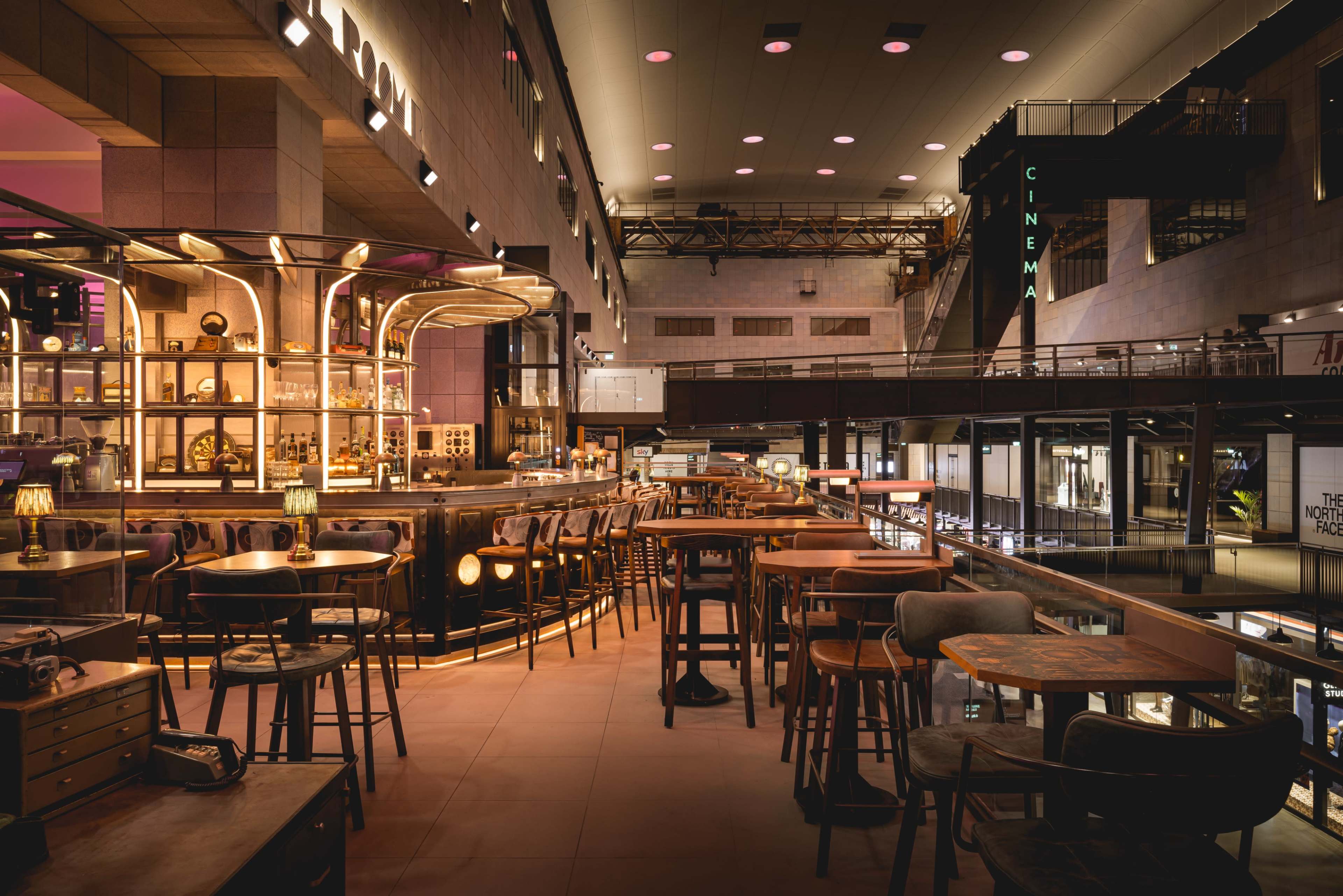 A modern restaurant interior featuring a bar area with illuminated shelves, numerous tables, and a view of an upper level with shops and a cinema sign.