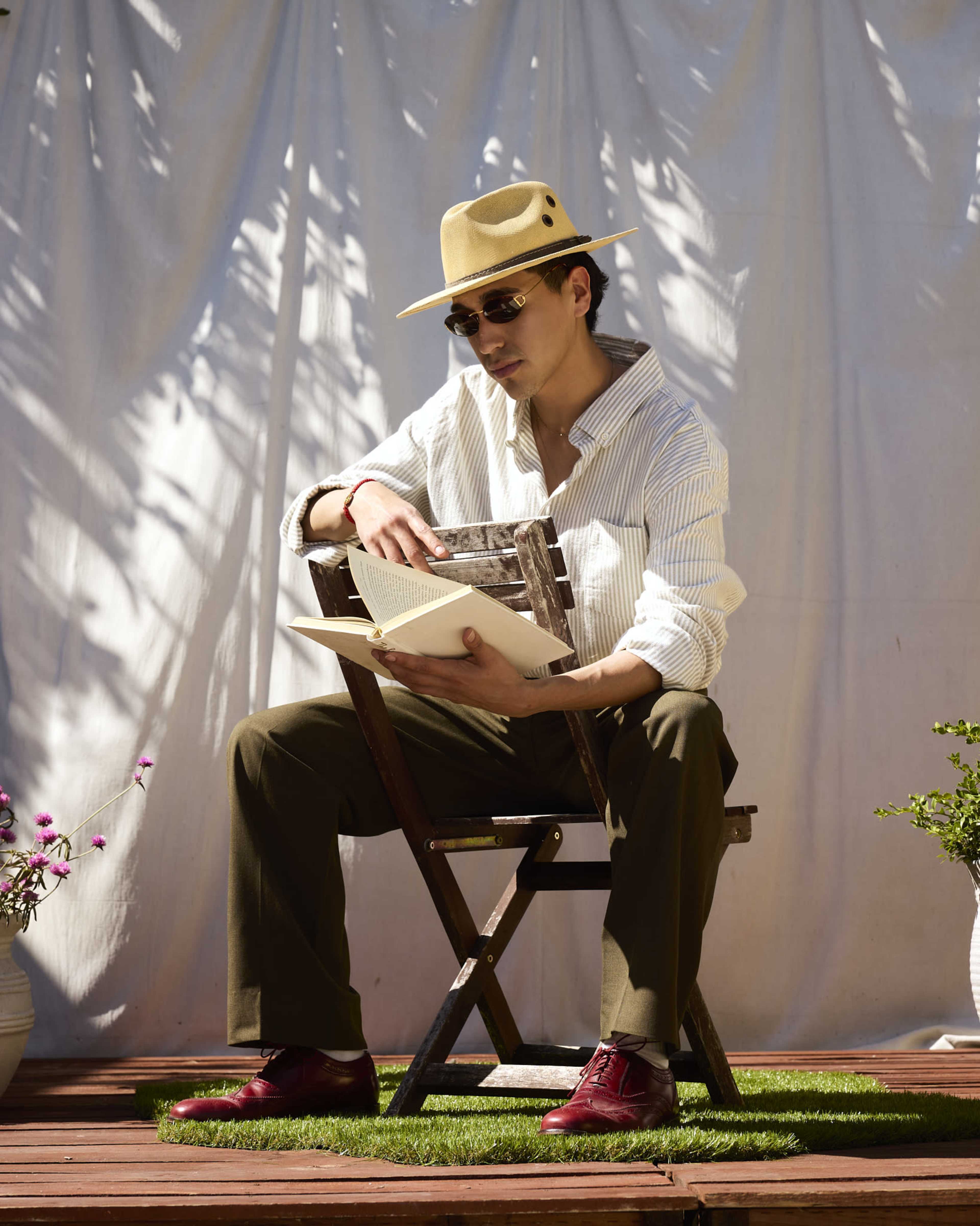 A person sits on a wooden chair, wearing a straw hat and sunglasses, reading a book in front of a white backdrop with potted plants nearby.