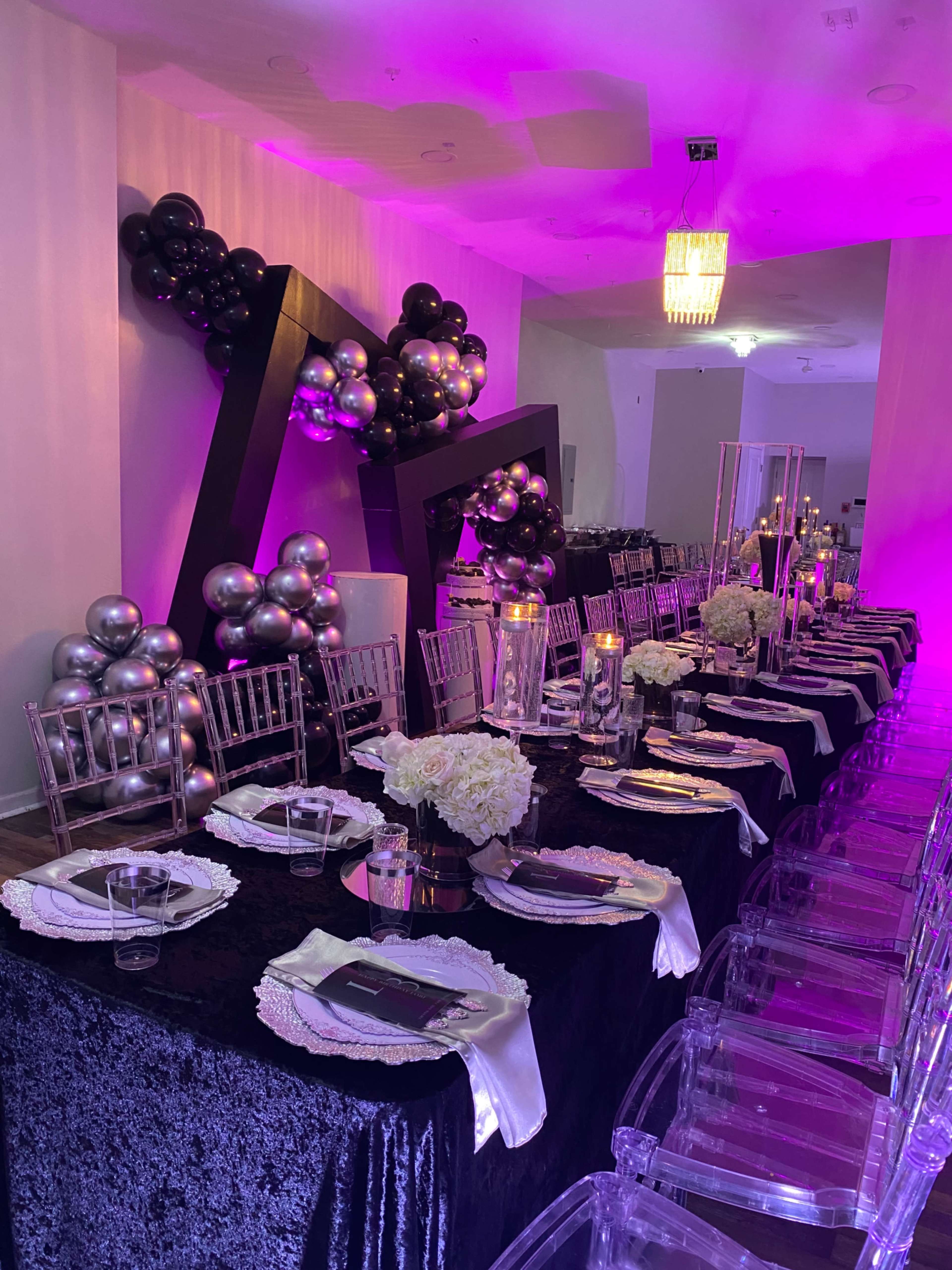A long dining table is set in a venue adorned with black, silver, and purple decorations, featuring clear acrylic chairs and floral centerpieces.