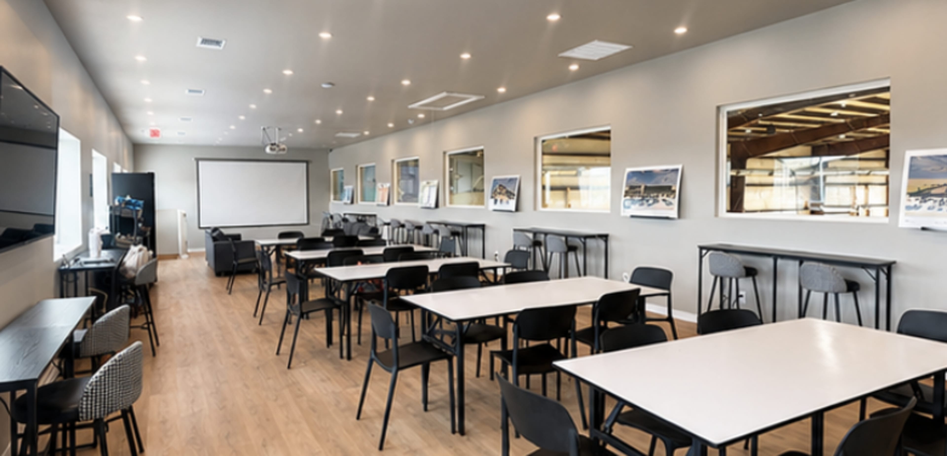 The image shows a spacious meeting room equipped with multiple tables and chairs, lines of windows, and a projector screen at one end.