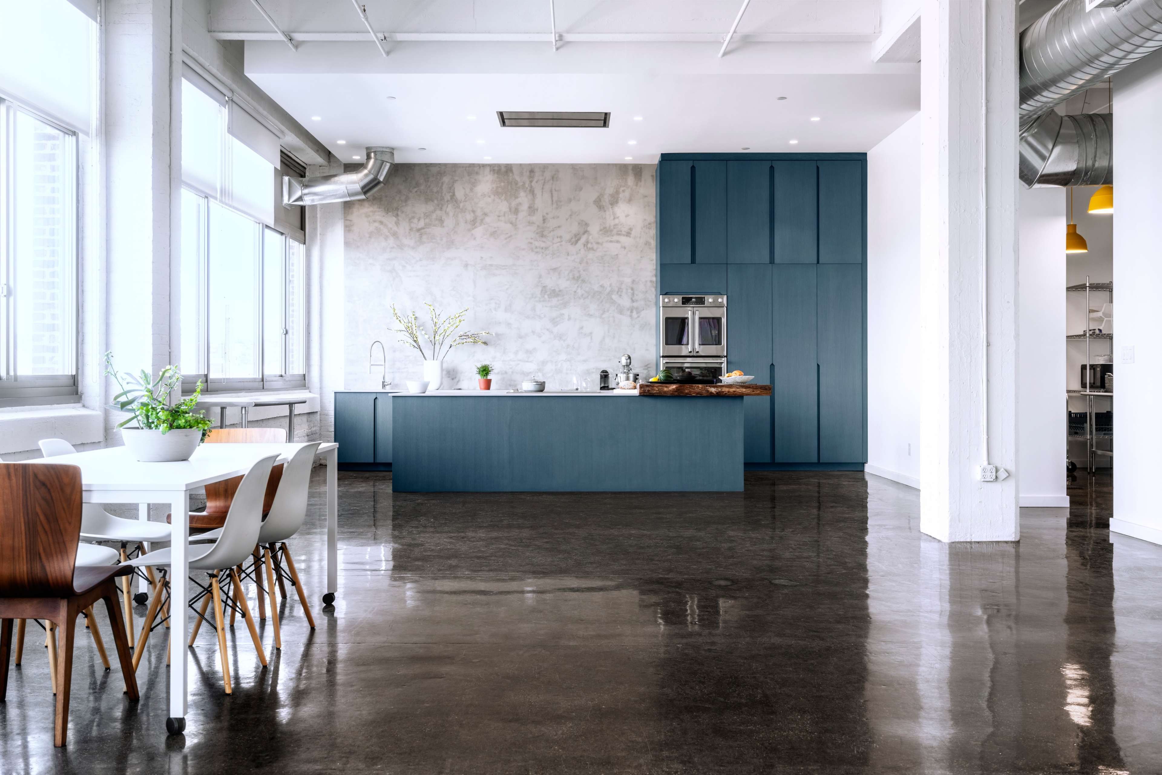 A modern kitchen features a blue island and cabinetry, with a dining area set nearby, and large windows allowing ample natural light.
