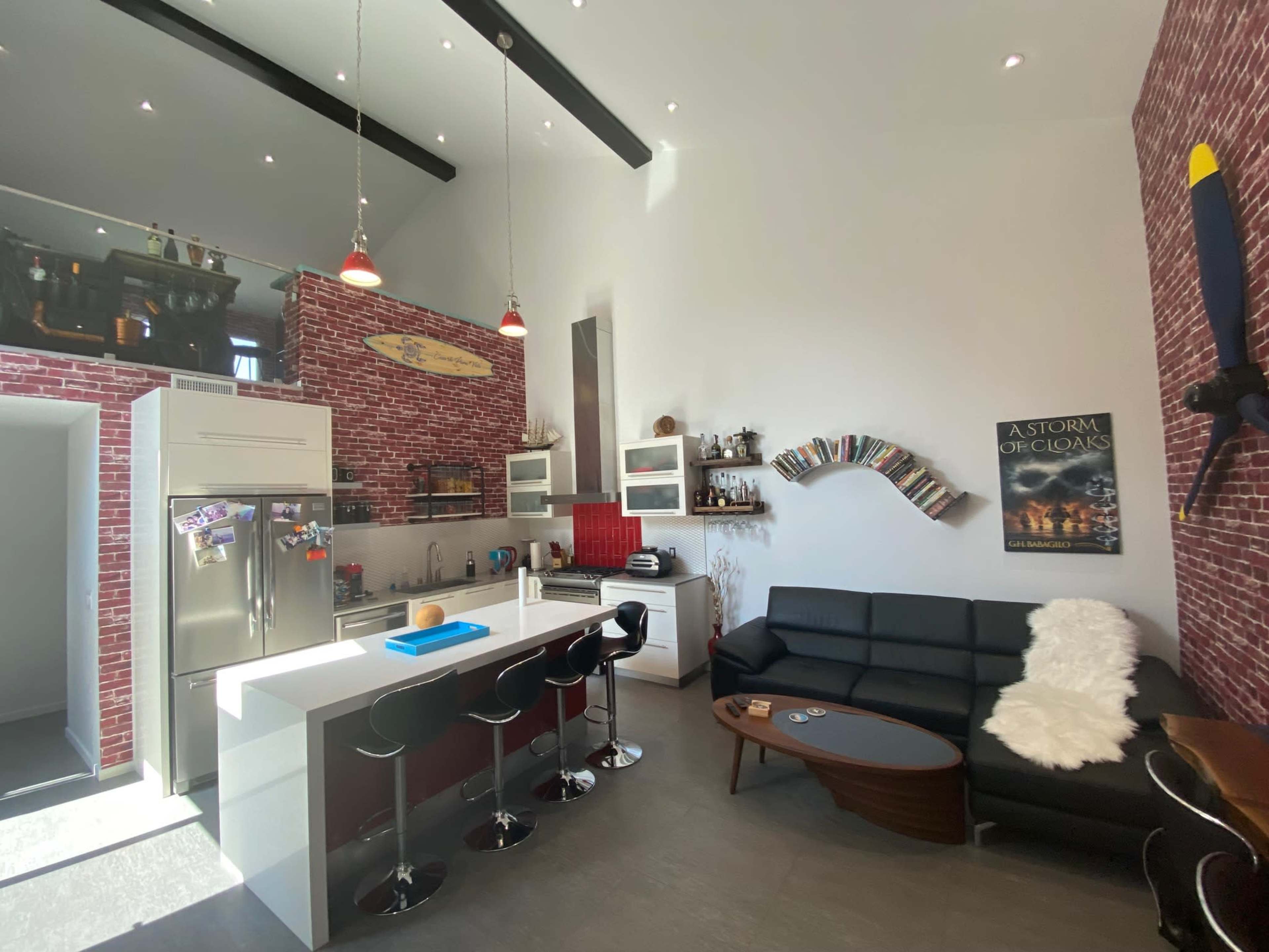 The image shows a modern kitchen with a high ceiling, featuring a brick wall, a large island with bar stools, and a seating area with a black couch and a coffee table.