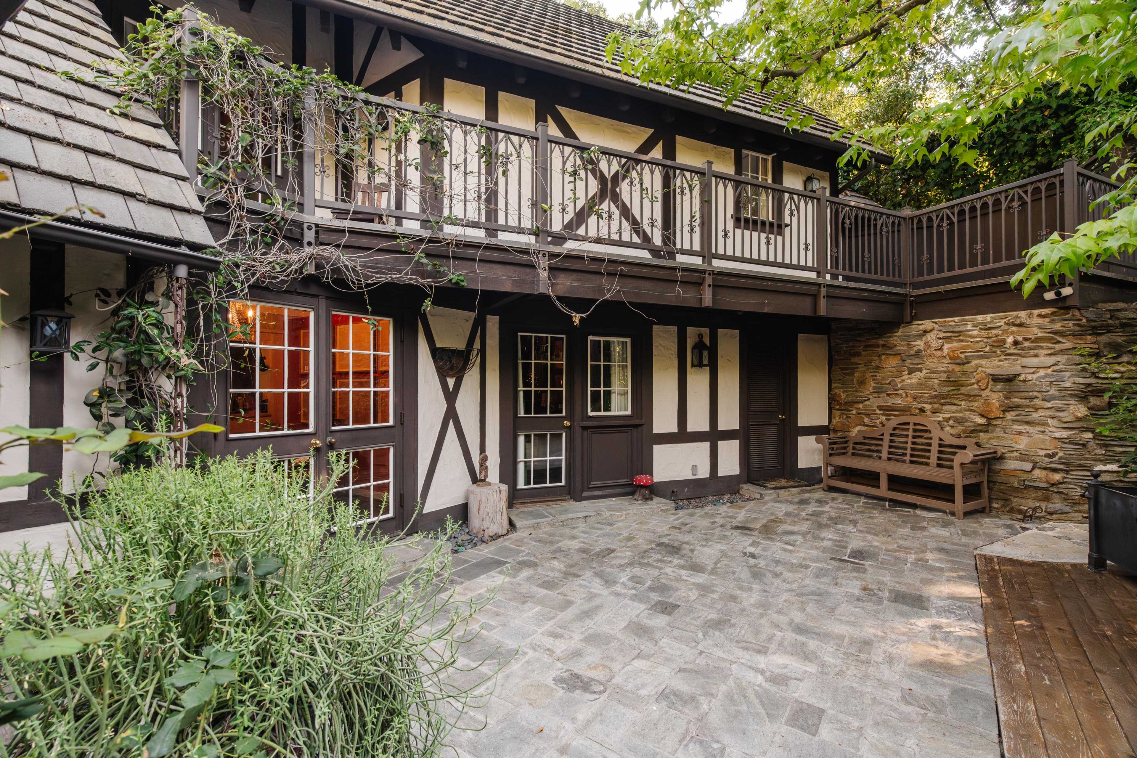 A stone-paved courtyard with a two-story timber-framed house featuring large windows and a balcony, surrounded by greenery.