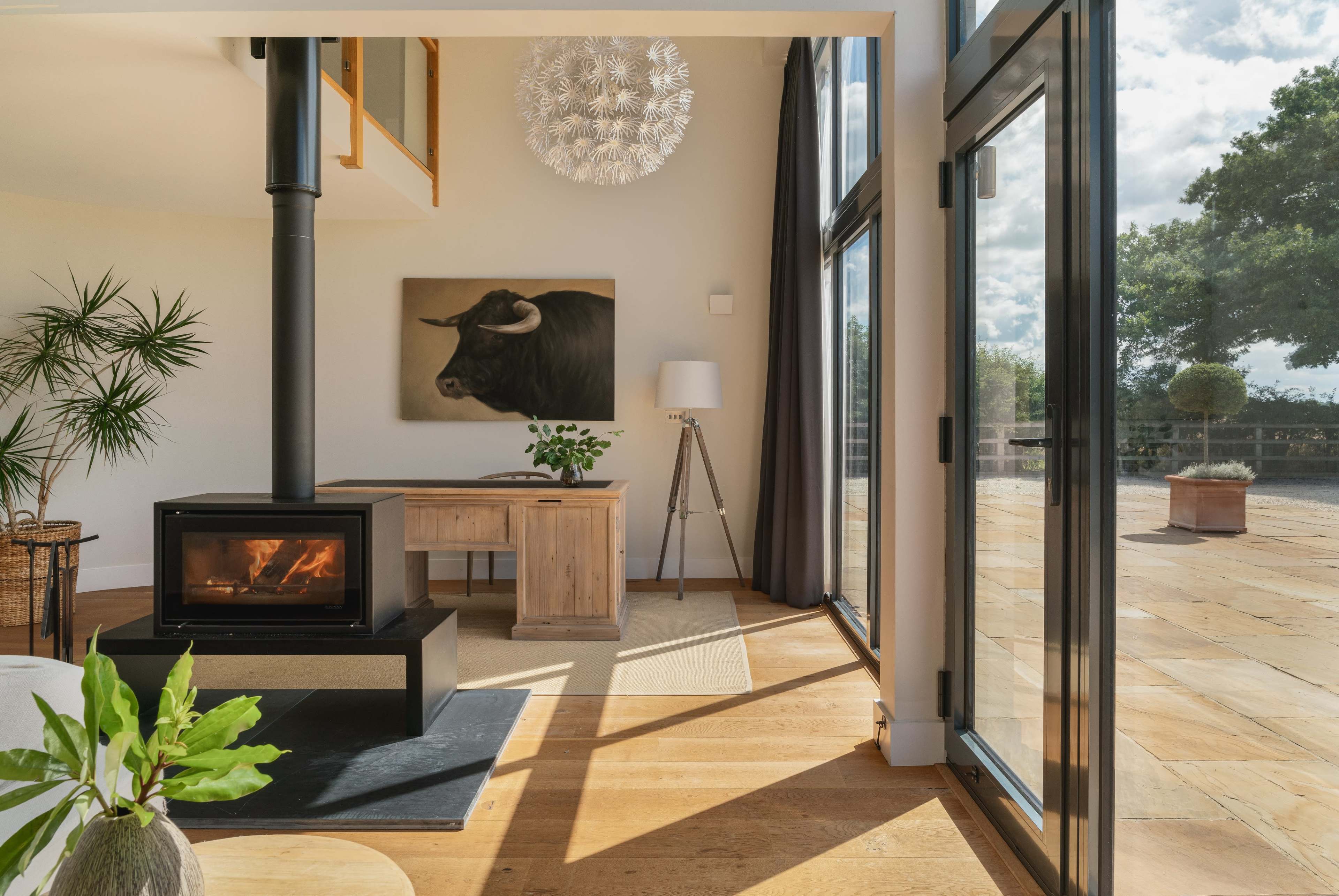A modern living space features a wood stove, a wooden desk, a large artwork of a bull on the wall, and large glass doors that open to a patio area.