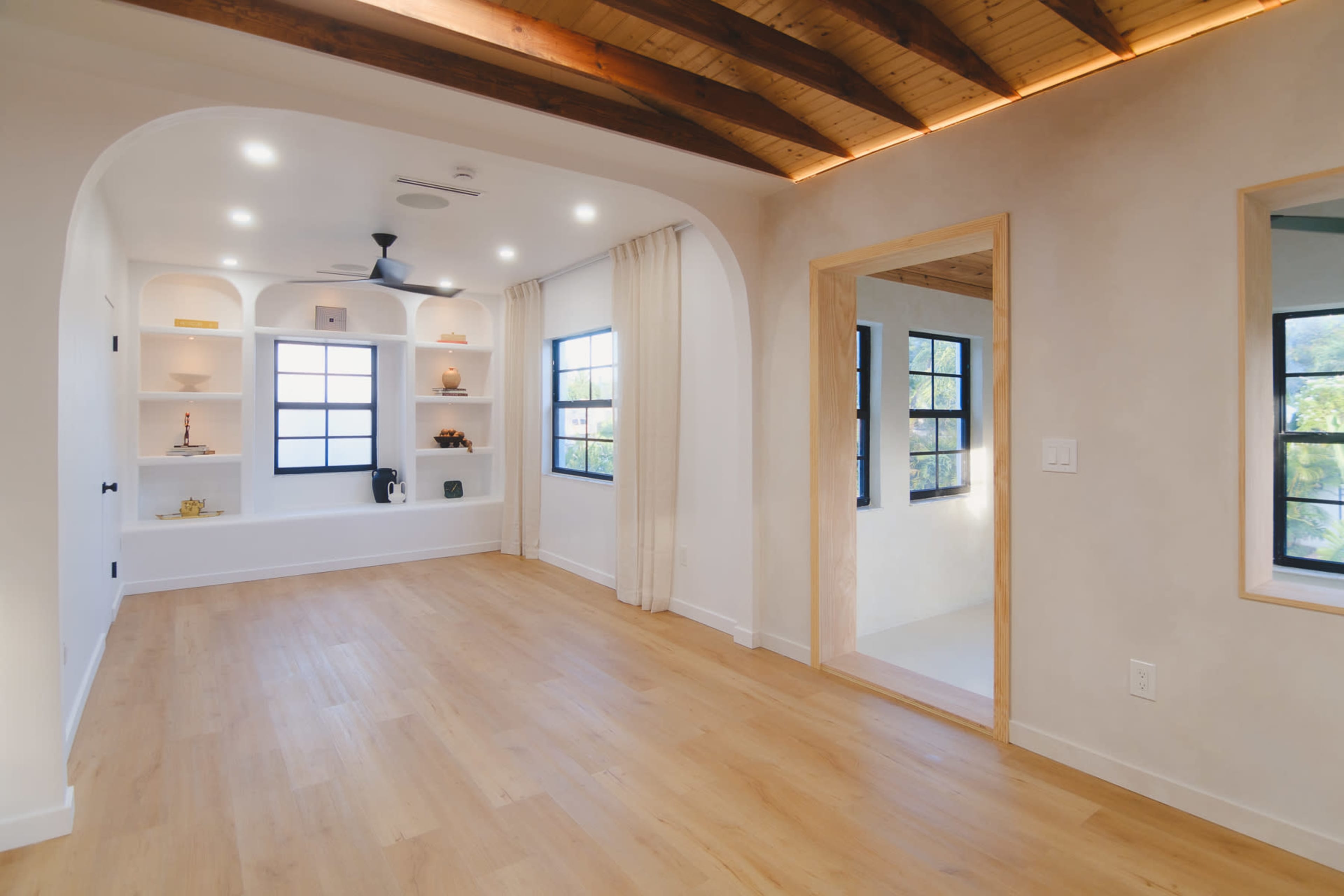 The image shows a bright, open room with wood-beamed ceilings, white walls, and shelves on one side filled with decorative items.