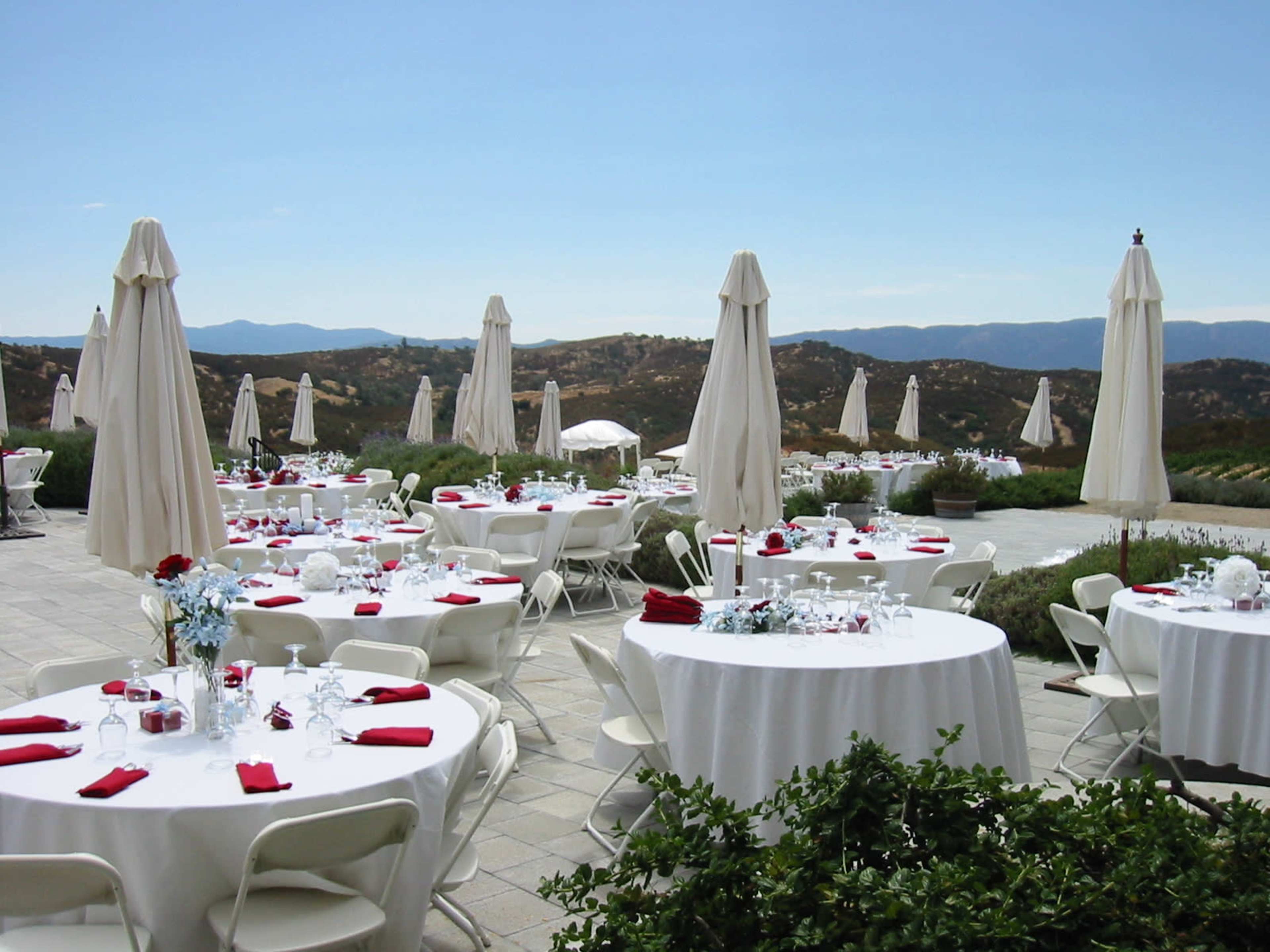 An outdoor event space features round tables set with white tablecloths and red napkins, surrounded by large umbrellas and overlooking a scenic landscape.