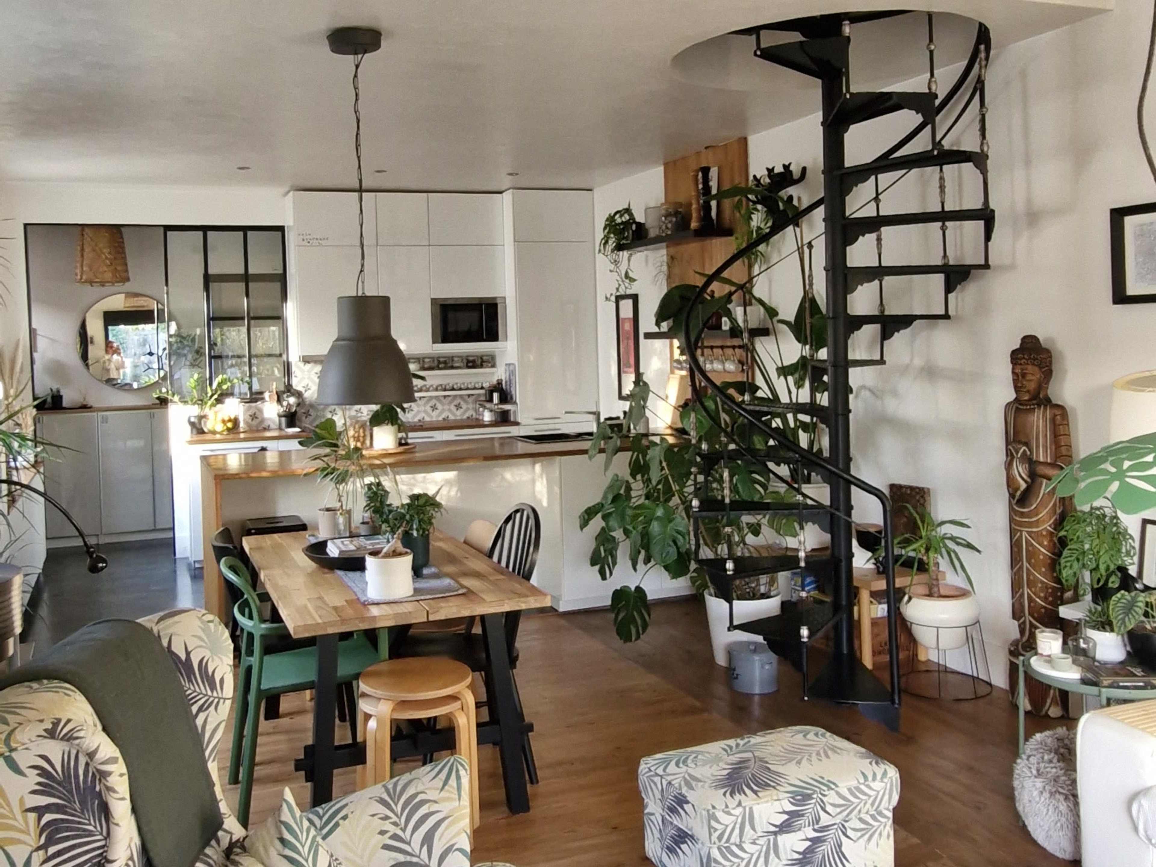 The image shows a modern open-concept living space featuring a spiral staircase, a dining area with wooden furniture, and various indoor plants.