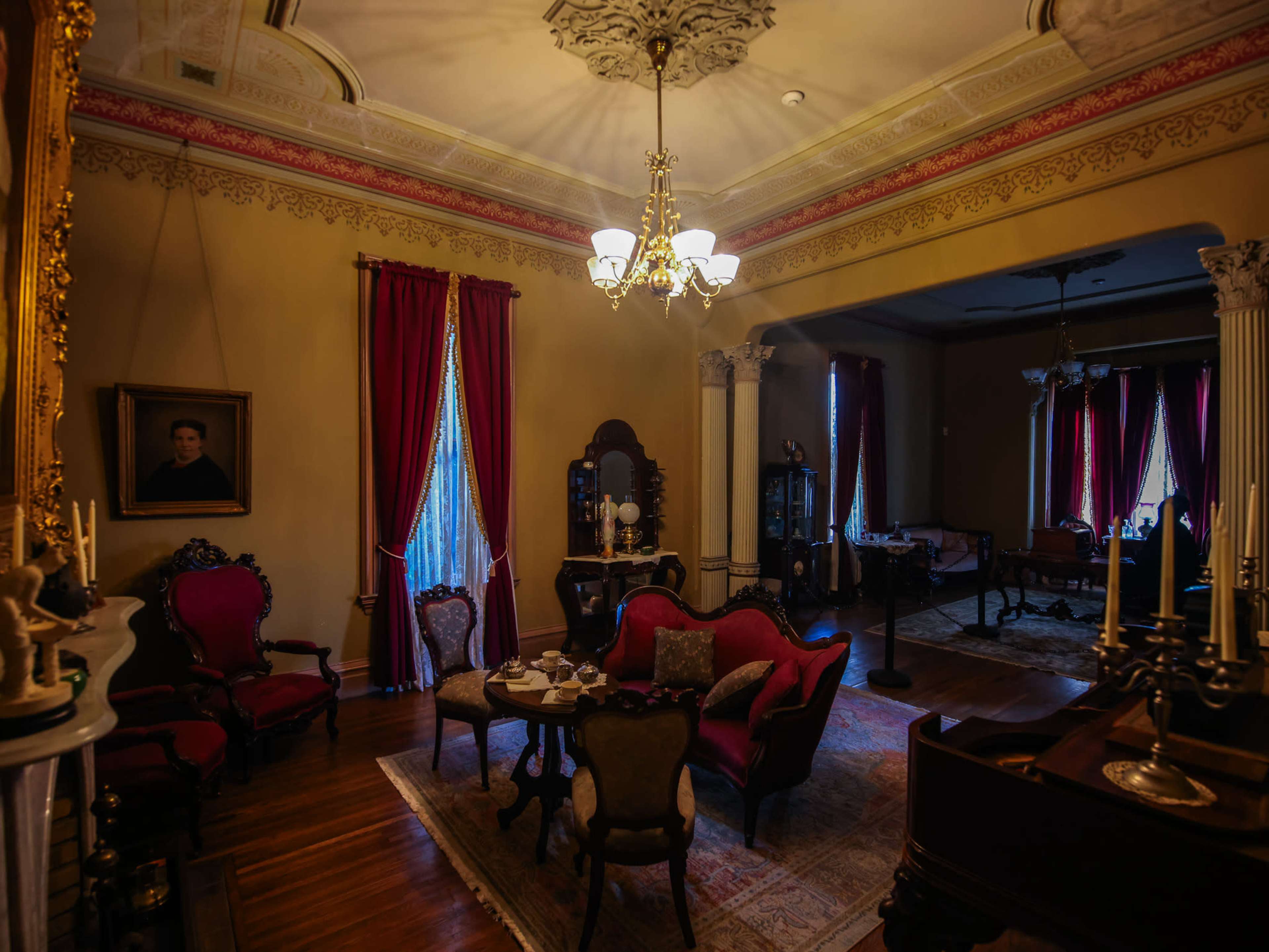 The room features vintage furnishings, including ornate chairs and a small table, with large red curtains framing the windows.
