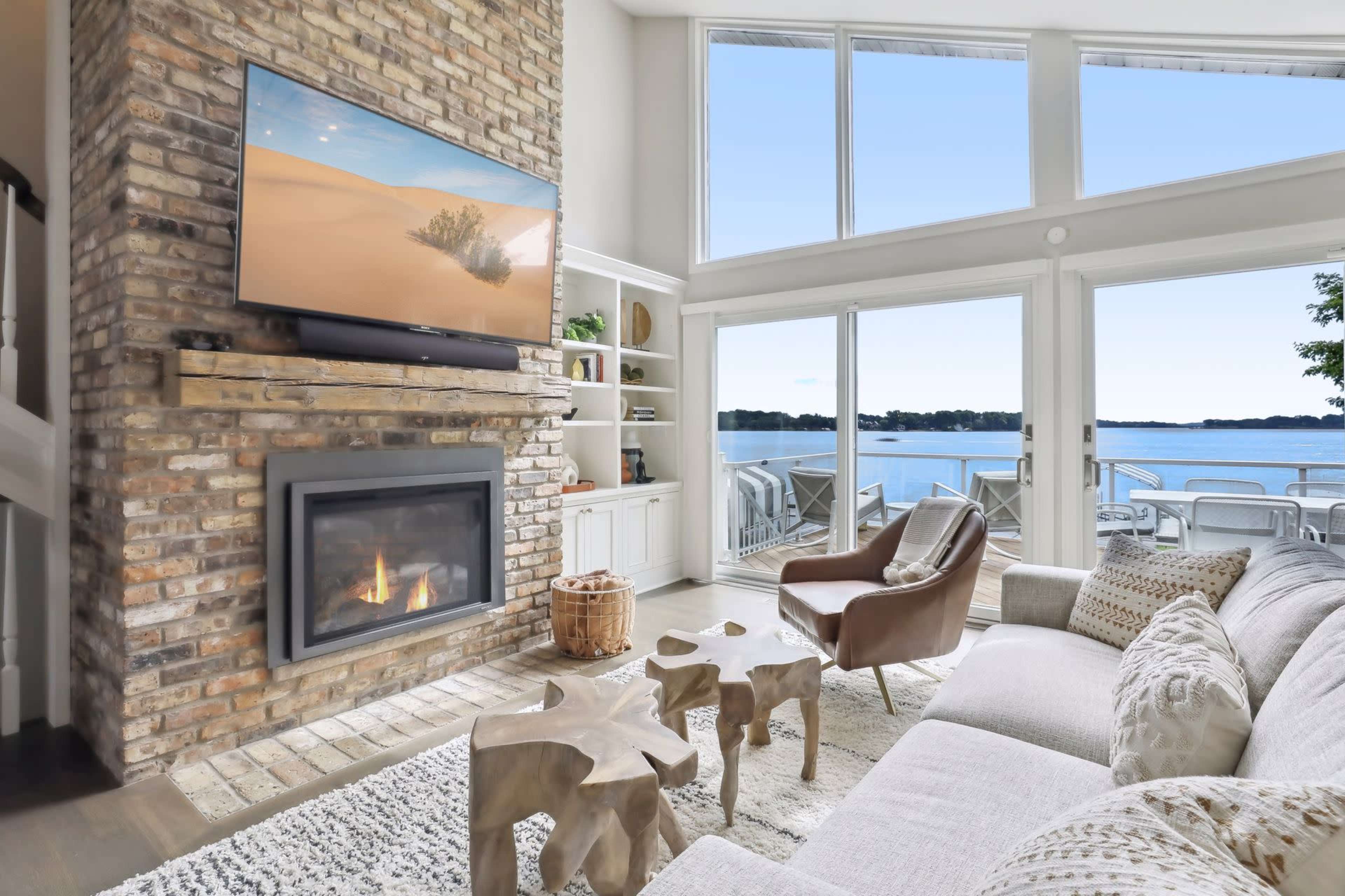 A modern living room with a brick wall, a large TV above a fireplace, and large windows overlooking a lake.