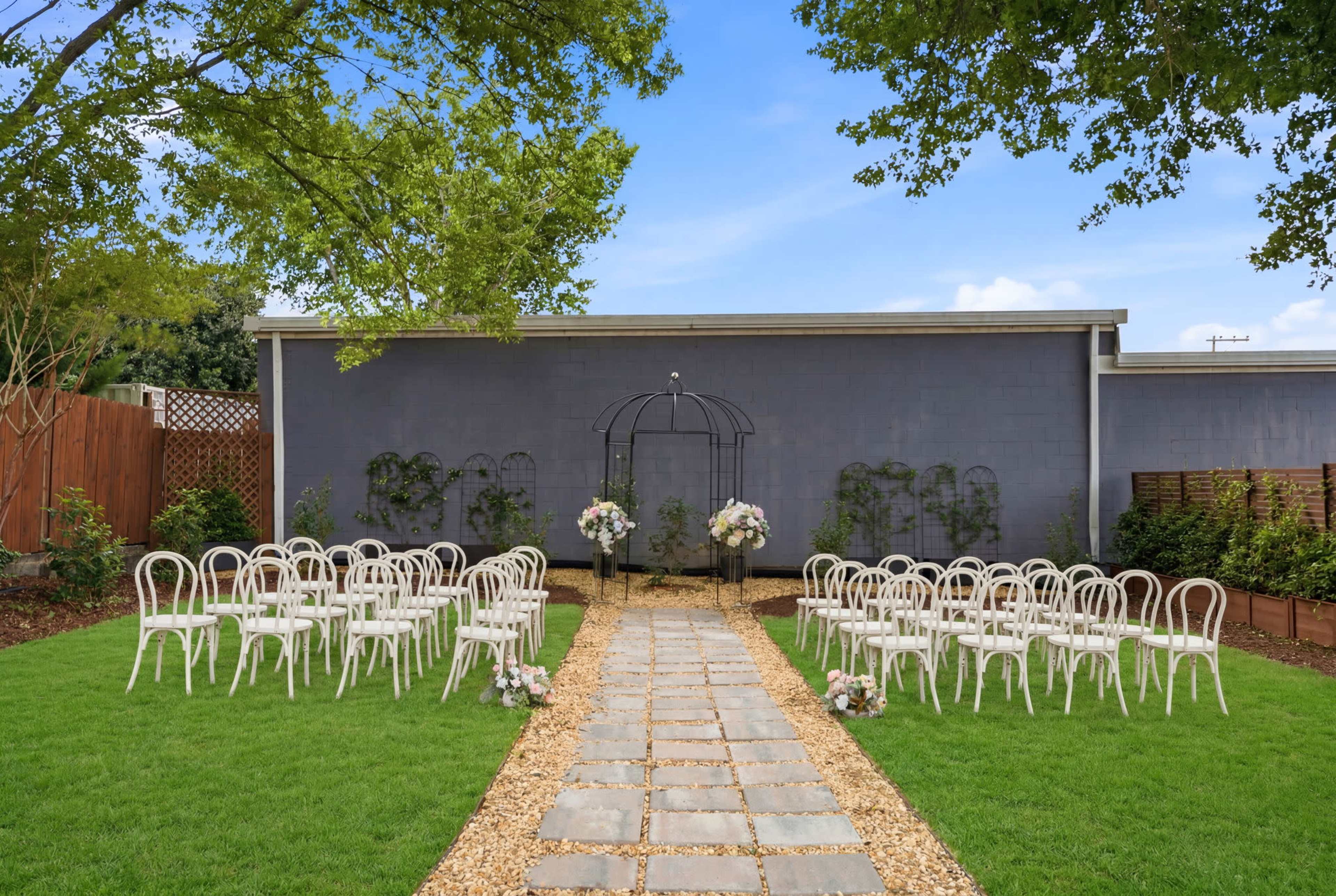 A neat outdoor wedding venue features a stone pathway leading to a floral-decorated arch, flanked by rows of white chairs on a grassy area.