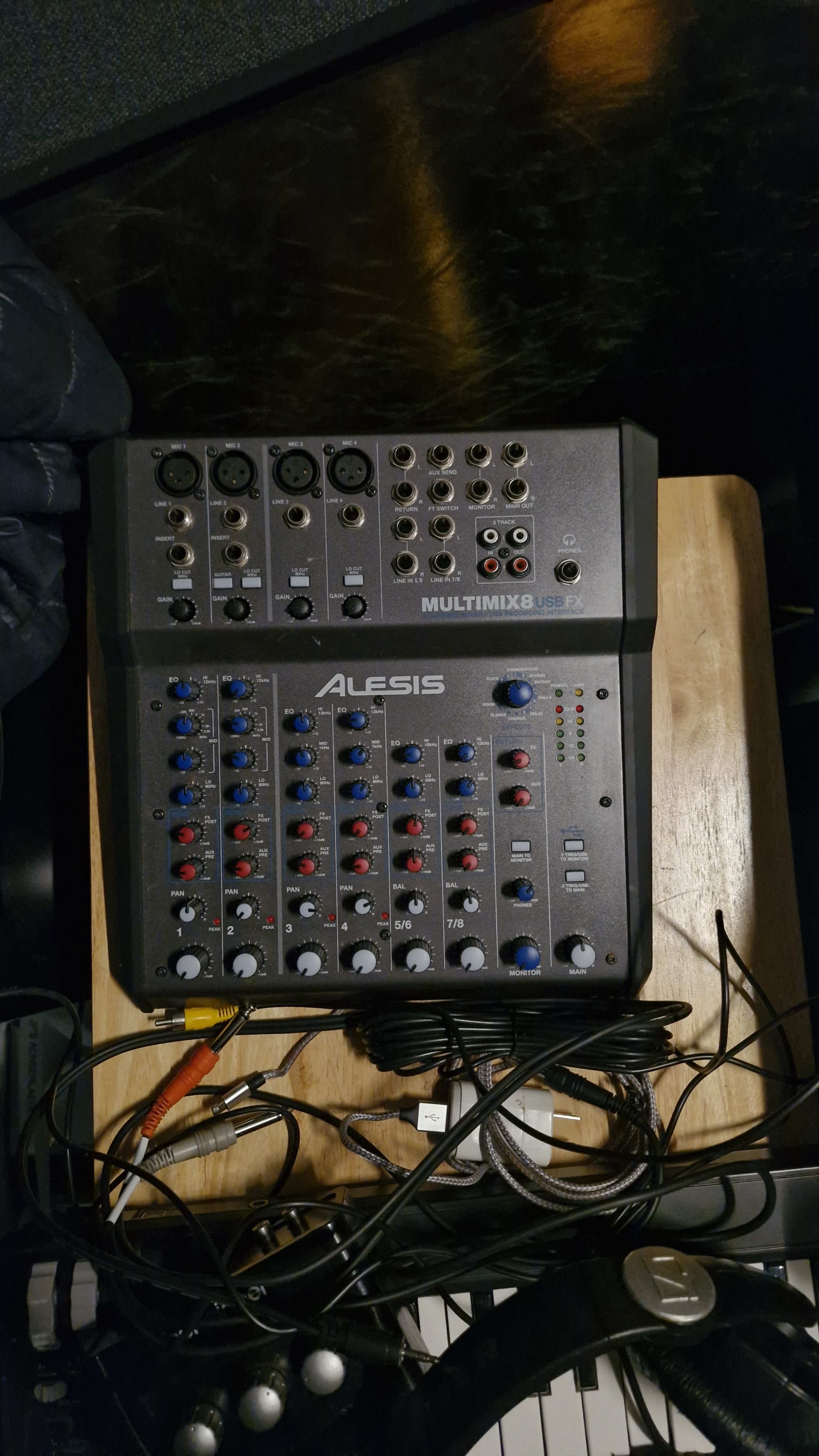 The image shows an Alesis MultiMix 8 USB audio mixer placed on a wooden surface, surrounded by various audio cables and a pair of headphones.