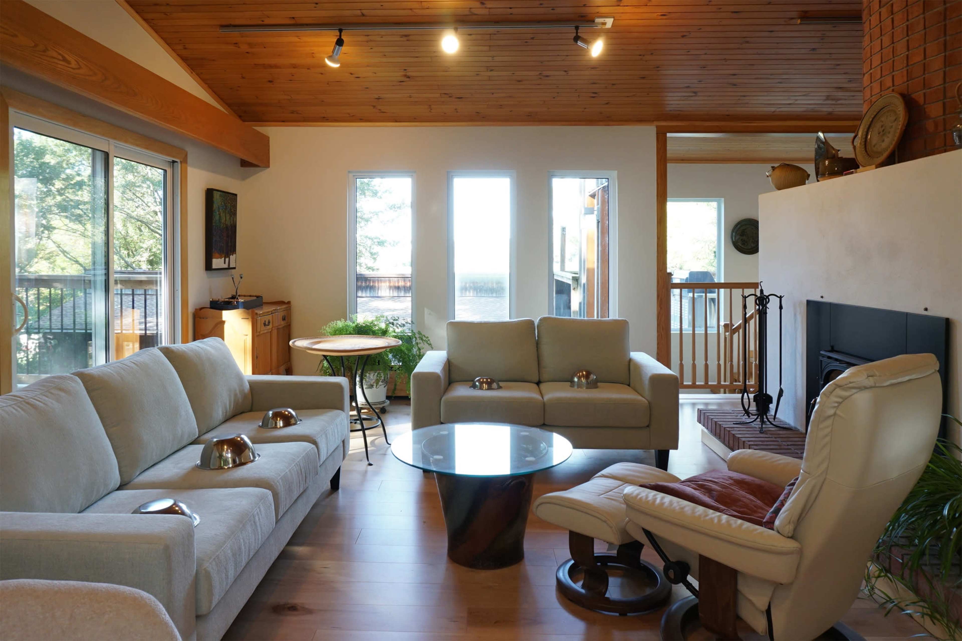 The image shows a modern living room with two light-colored sofas, a round glass coffee table, and a comfortable armchair near a fireplace, surrounded by large windows.