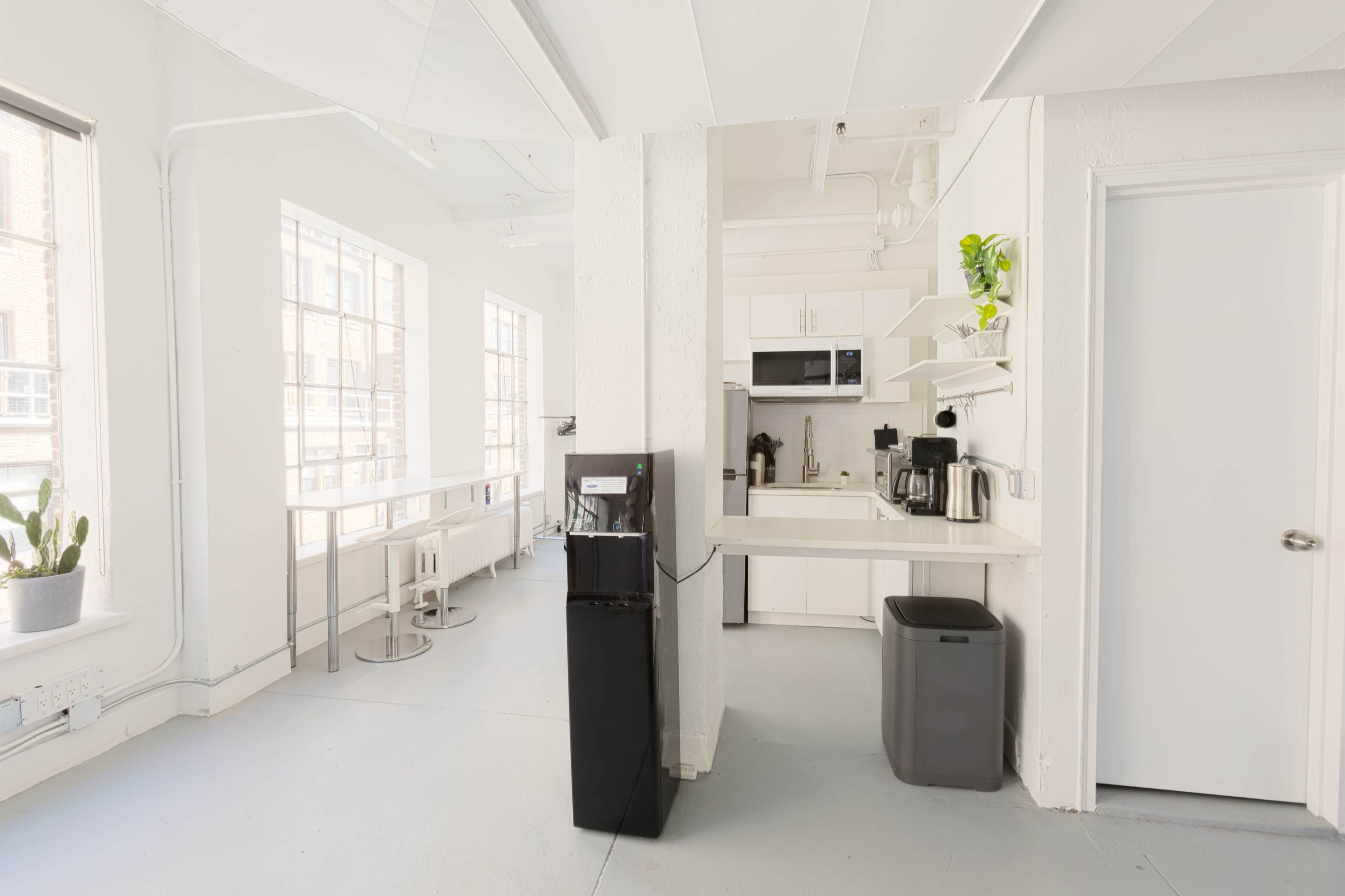 A bright, minimalist kitchen area featuring a water cooler, coffee station, and a dining space with tall windows and a white color scheme.